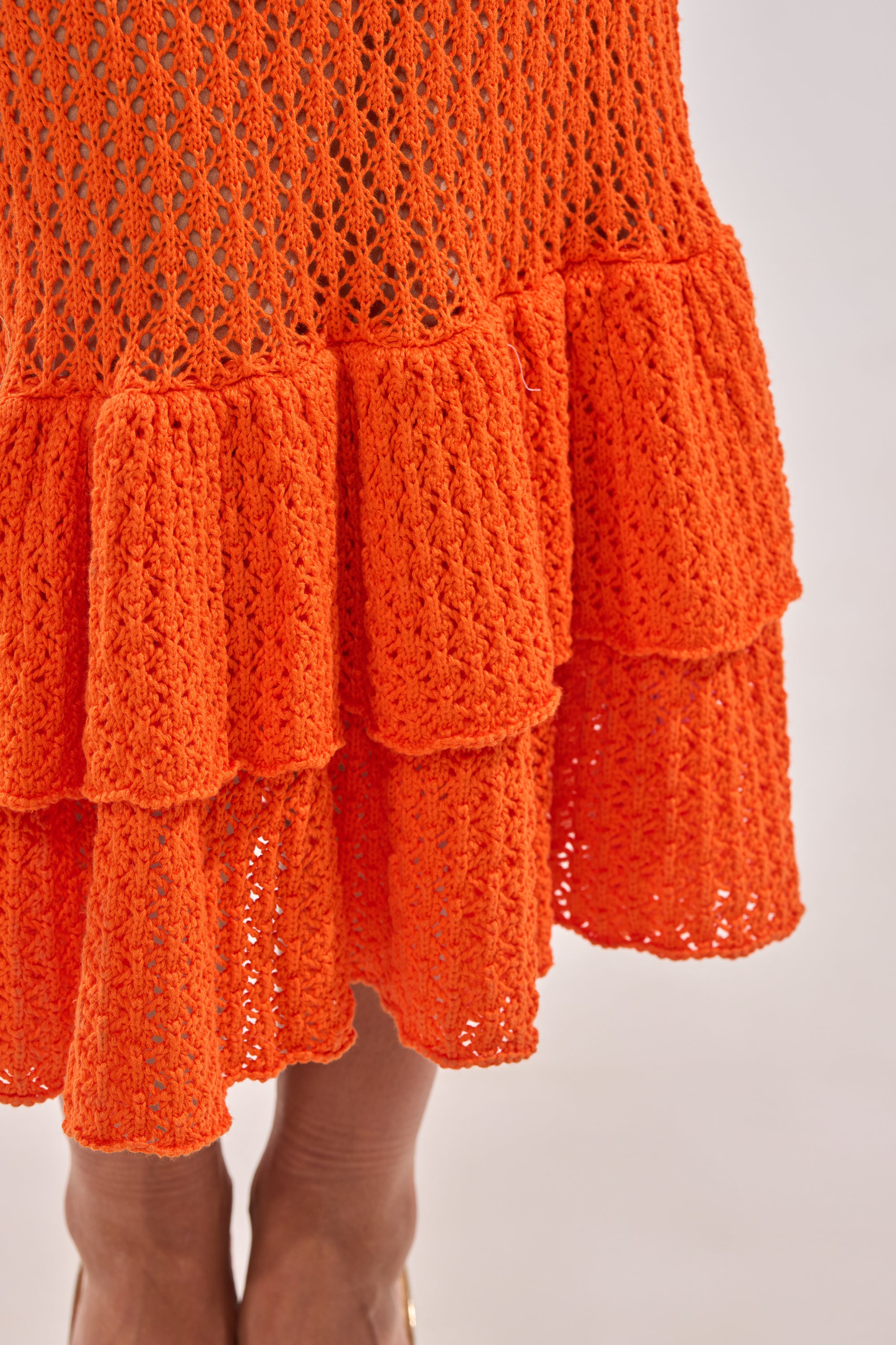 Close-up of the lower half of the LINSEY MAXI KNIT DRESS IN ORANGE, featuring intricate crochet patterns and a ruffled hem, worn by a person against a plain background. 5