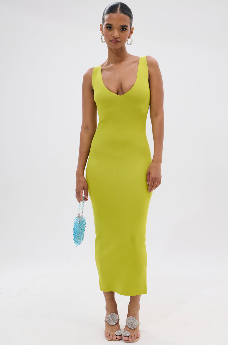 Against a white background, a woman wears the BETWEEN THE LINES MIDI DRESS IN LIME—a sleeveless, fitted summer staple—styled with a blue beaded purse, pulled-back hair, hoop earrings, and flat sandals featuring decorative bows. 1