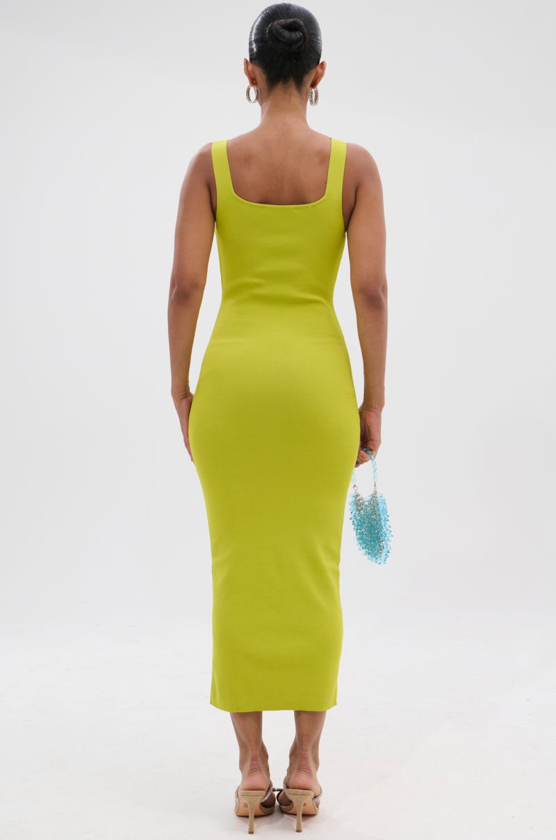 A woman faces away in the BETWEEN THE LINES MIDI DRESS IN LIME, a fitted sleeveless summer staple, paired with beige heels and a blue beaded bag. Her dark hair is in a neat bun, set against a plain white background. 3