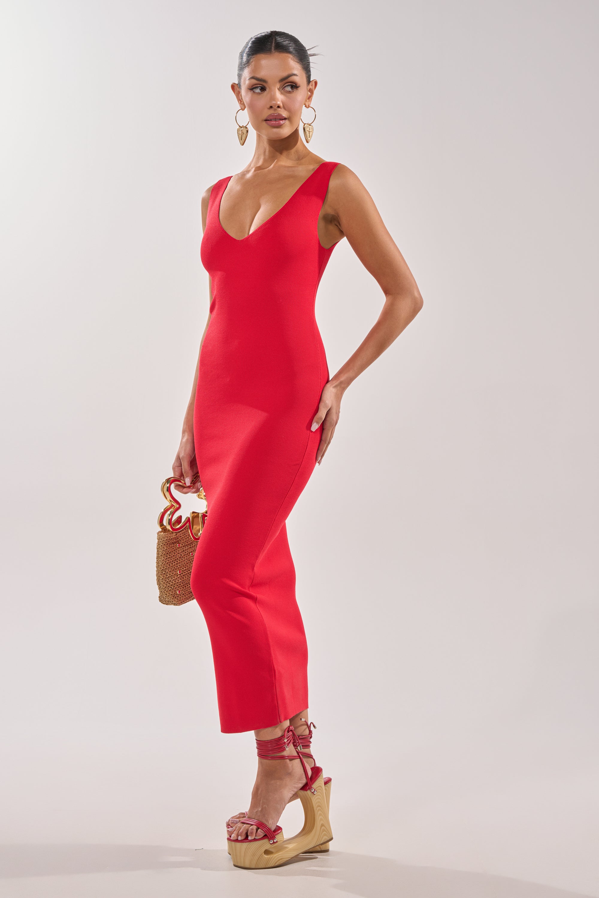 A woman stands against a plain light background wearing the BETWEEN THE LINES MIDI DRESS IN RED with a plunging neckline, red platform sandals with ankle straps, and holding a small woven handbag. 2