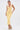 A woman stands against a white background in the BETWEEN THE LINES MIDI DRESS IN YELLOW from AKIRA Label, a sleeveless V-neck style, paired with colorful platform sandals and large yellow flower earrings, her hair pulled back.