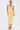 A woman wearing the BETWEEN THE LINES MIDI DRESS IN YELLOW—a fitted, sleeveless midi with a deep V-neckline—pairs it with colorful flower earrings and rainbow-striped platform sandals against a plain white background.