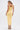 A woman with her hair tied back and round earrings models the BETWEEN THE LINES MIDI DRESS IN YELLOW, paired with colorful platform heels, against a plain white background.