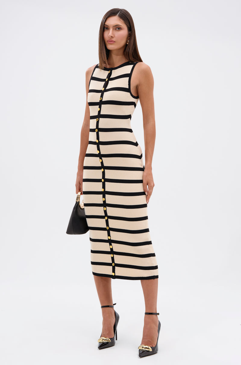A woman poses against a white background wearing the KAREN STRIPE KNIT MIDI DRESS with black horizontal stripes, gold buttons down the front, and stretchy knit fabric. She accessorizes with a black handbag and black heels featuring gold accents.