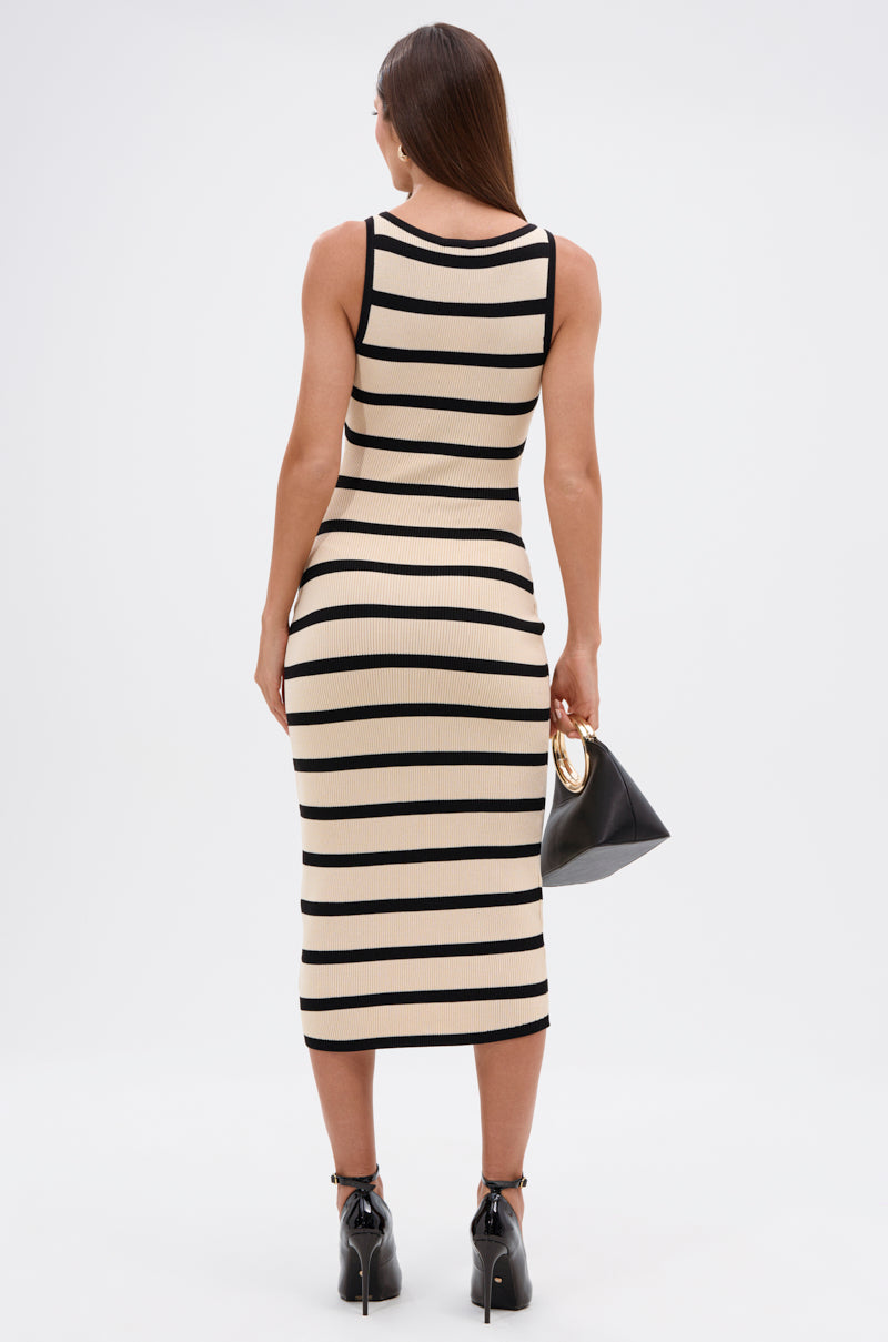 A woman faces away, wearing the KAREN STRIPE KNIT MIDI DRESS with wide black horizontal stripes. She carries a black handbag with a gold handle and pairs the dress with black heeled ankle boots.
