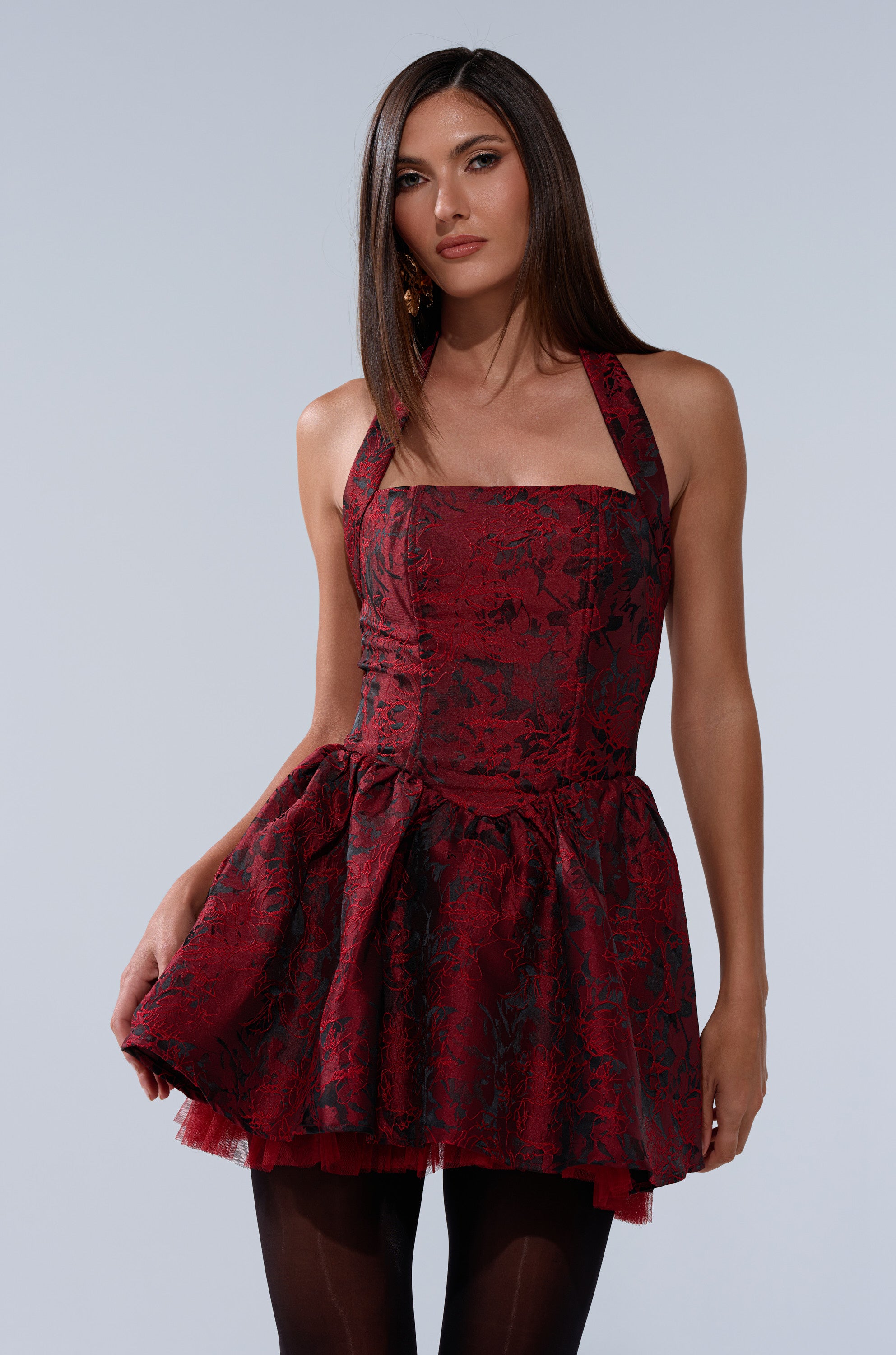 A woman with long straight brown hair wears the BRINA BROCADE MINI DRESS IN BURGUNDY, a sleeveless flared dress paired with black tights, standing against a plain light gray background.