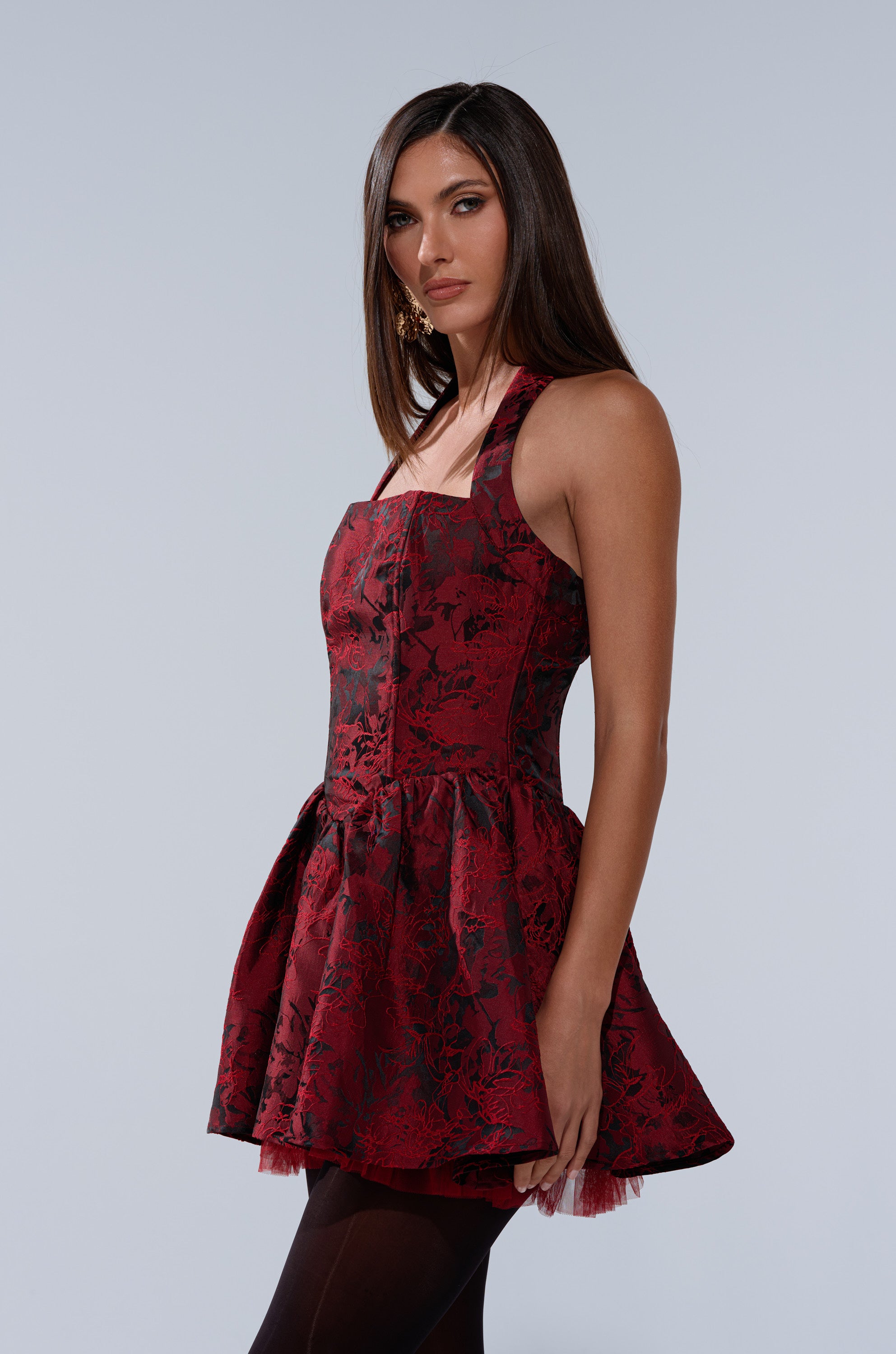A woman with long, straight brown hair wears the BRINA BROCADE MINI DRESS IN BURGUNDY, a sleeveless mini dress with a skater silhouette and flared skirt. She stands against a light gray background, gazing slightly to the side.