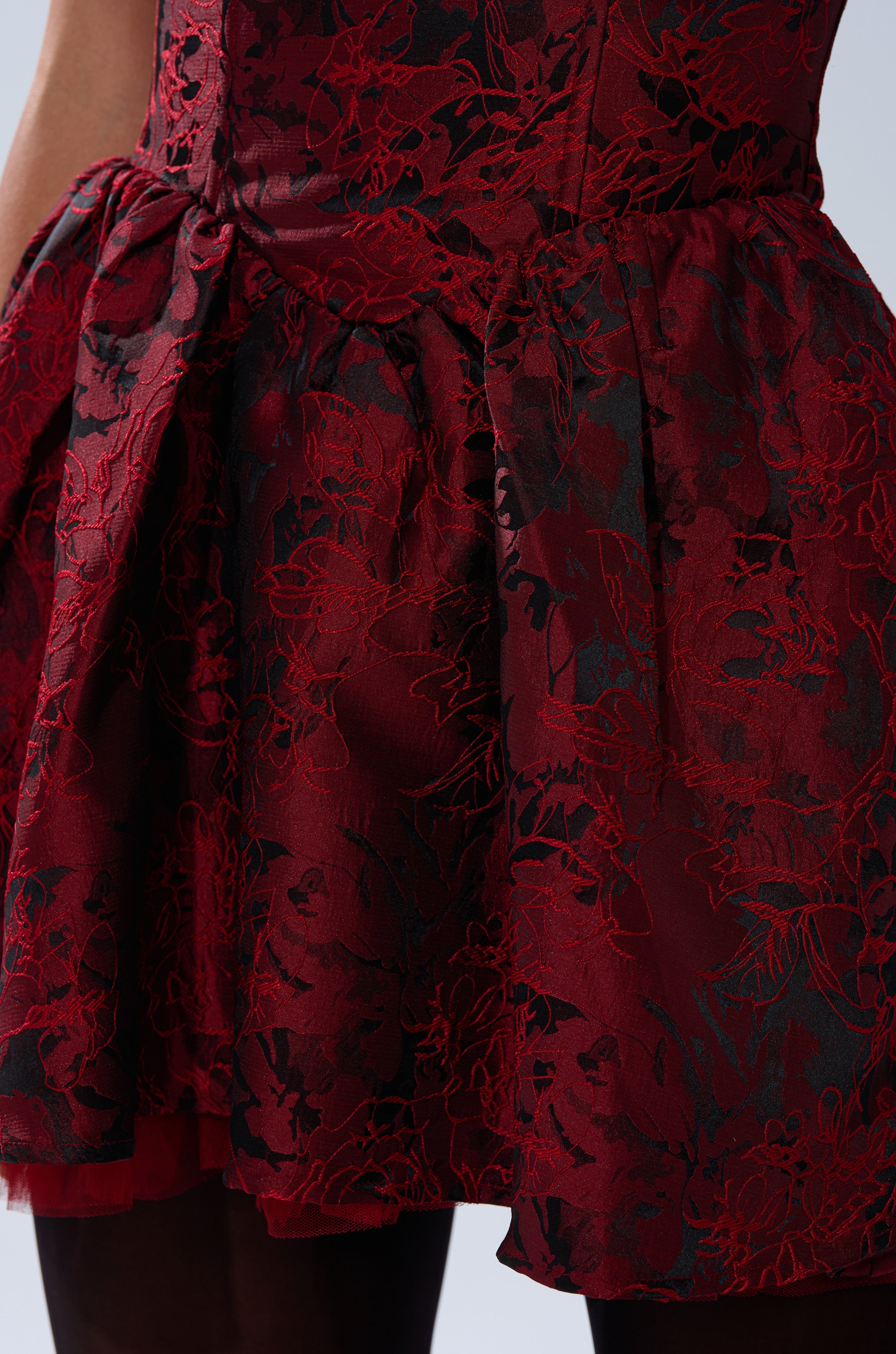A close-up shows someone wearing the BRINA BROCADE MINI DRESS IN BURGUNDY, featuring a flared skirt, fitted waist, and intricate lace details. Part of their arm and stockings are also visible.