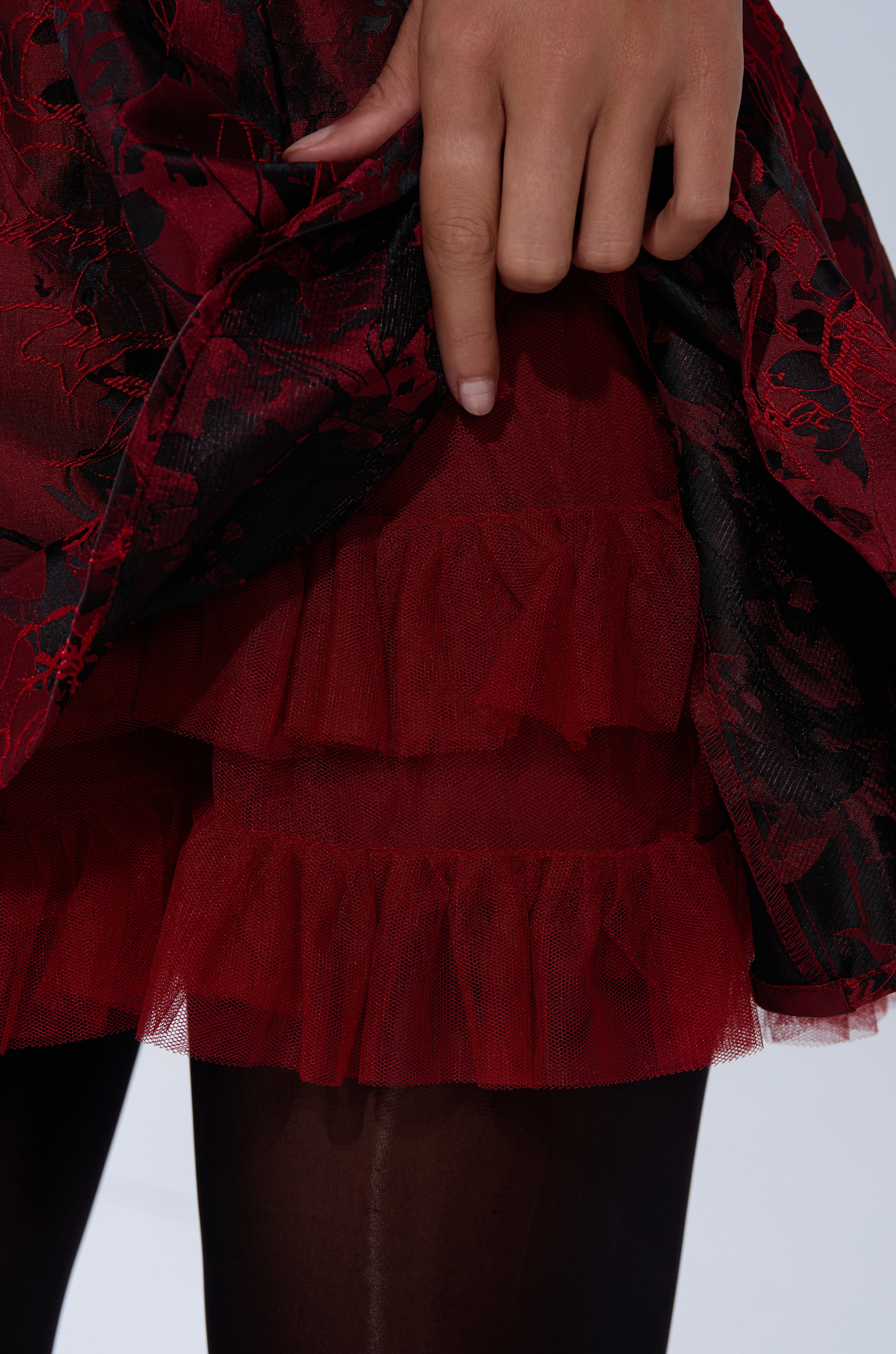A close-up of a person lifting the BRINA BROCADE MINI DRESS IN BURGUNDY to reveal layers of red ruffled tulle underneath, paired with black tights.