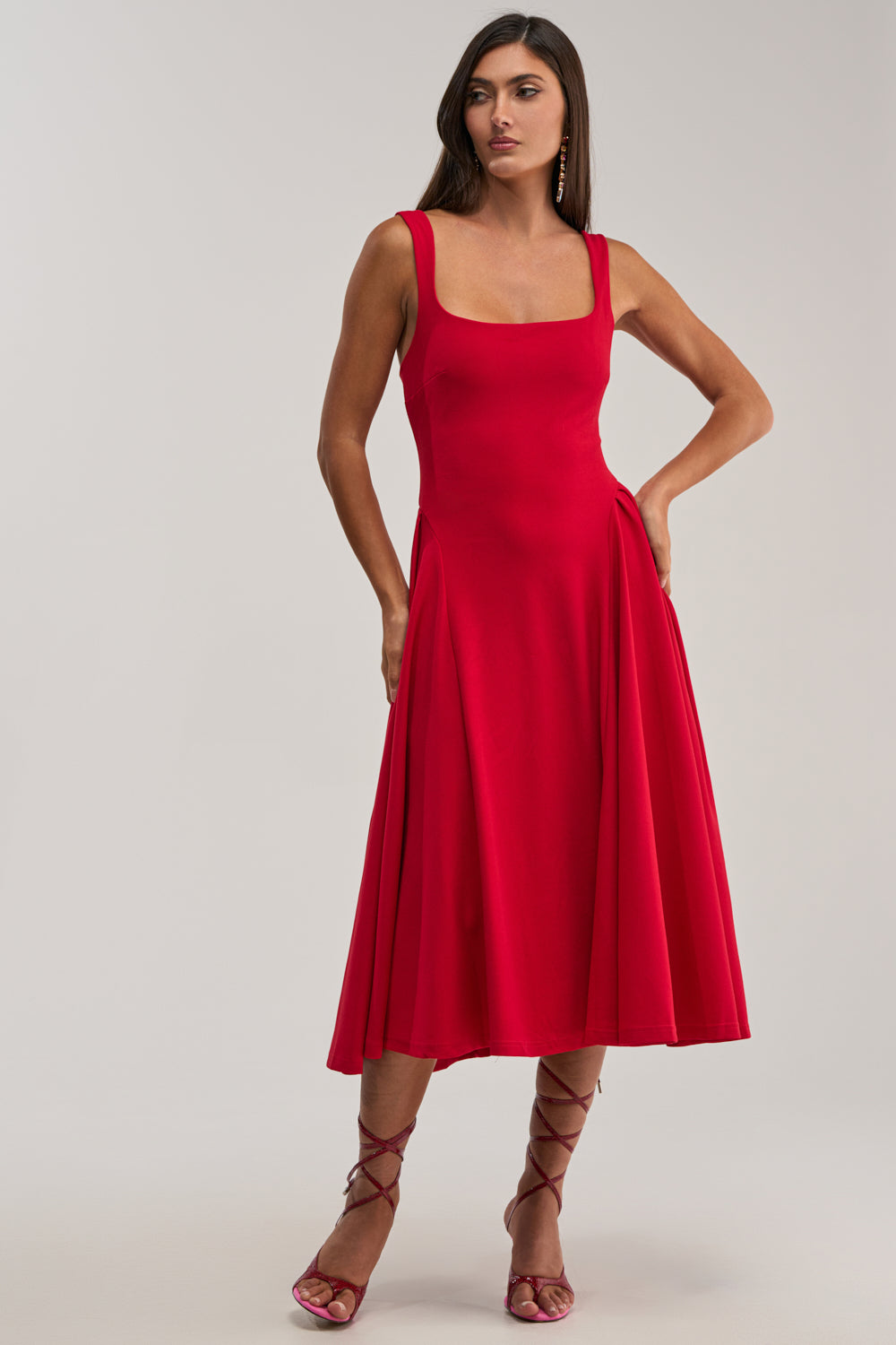 A woman stands with one hand on her hip, wearing the BOSS BABE MIDI DRESS—a sleeveless red fit-and-flare midi dress with a square neckline—paired with strappy red heels against a plain light background.
