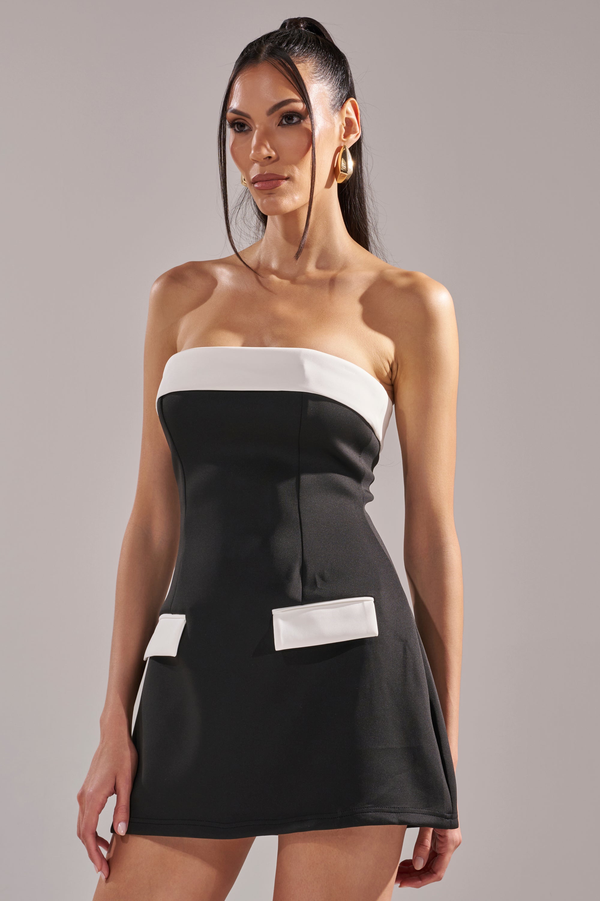 A woman wears the TOO CUTE MINI DRESS, a strapless color-block mini dress featuring a white band across the top and two white decorative flaps at the hips, standing against a plain gray background.