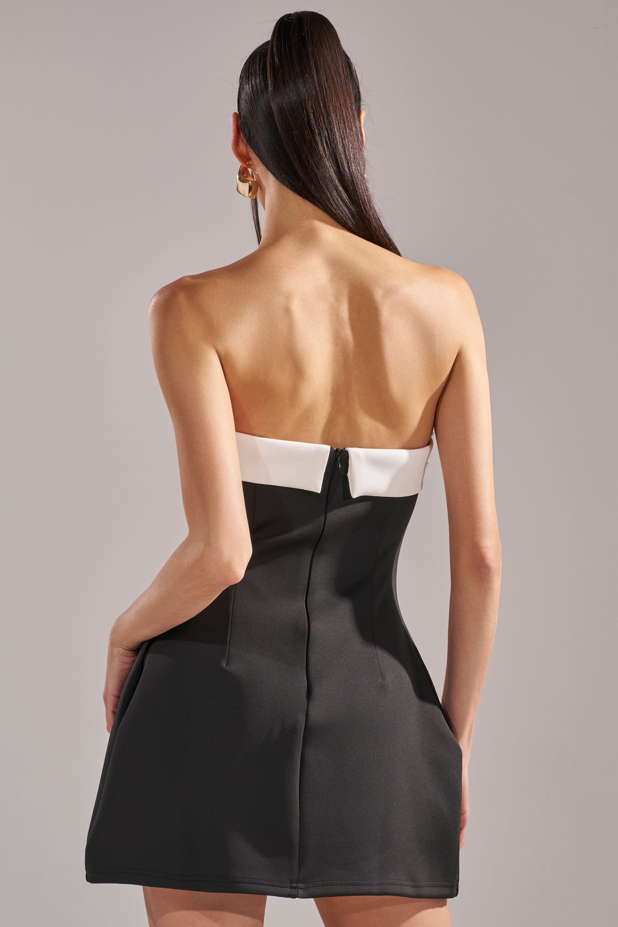 A woman with long, straight dark hair models the TOO CUTE MINI DRESS—a black and white strapless mini with a white top band, back zipper, and fitted color-block silhouette—standing against a plain background.