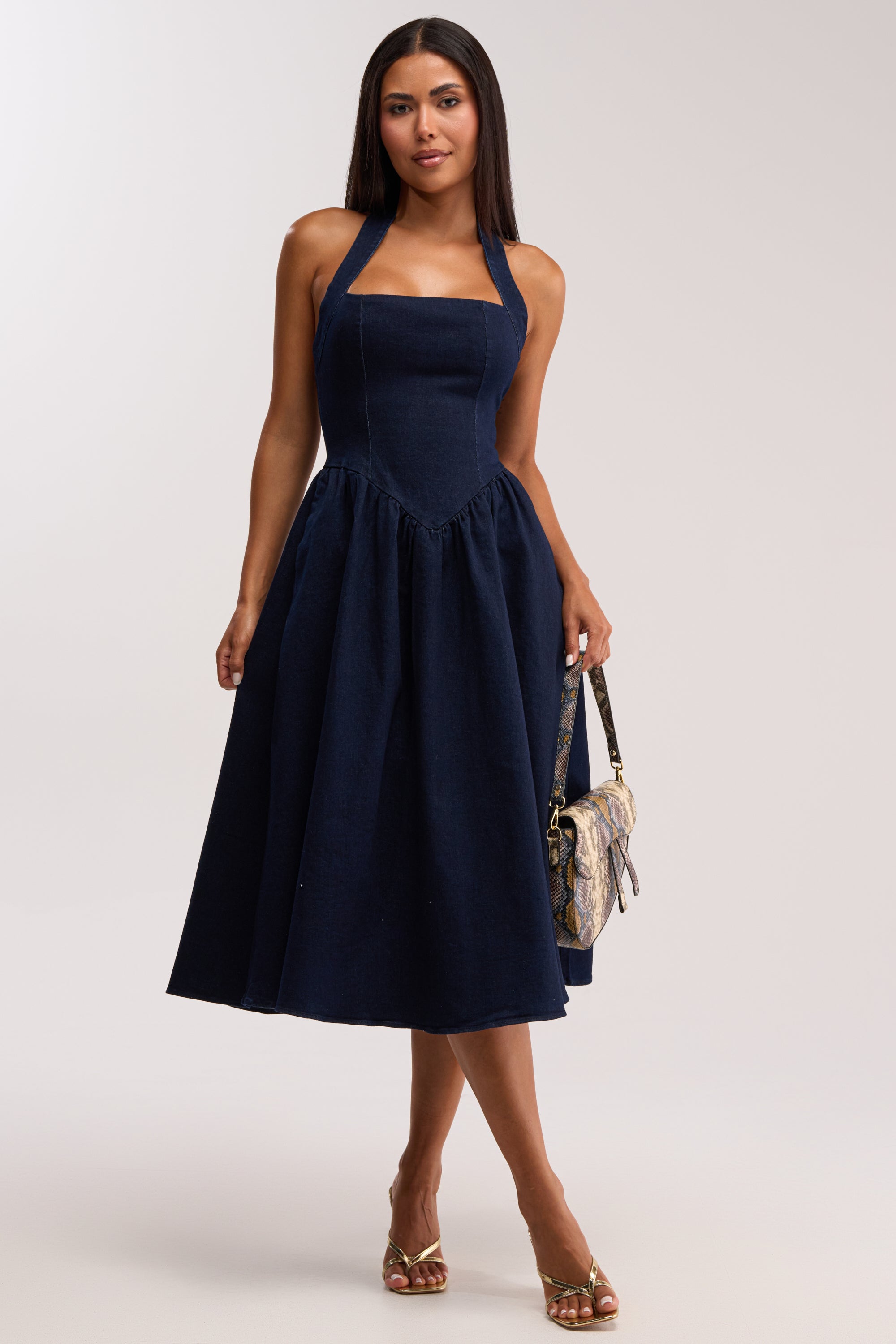 A woman wearing the MISS INDEPENDENT DENIM HALTER MIDI DRESS, featuring a sleeveless navy blue design with a fitted bodice and flared skirt, stands holding a patterned handbag. She pairs gold sandals with her straight, shoulder-length hair.