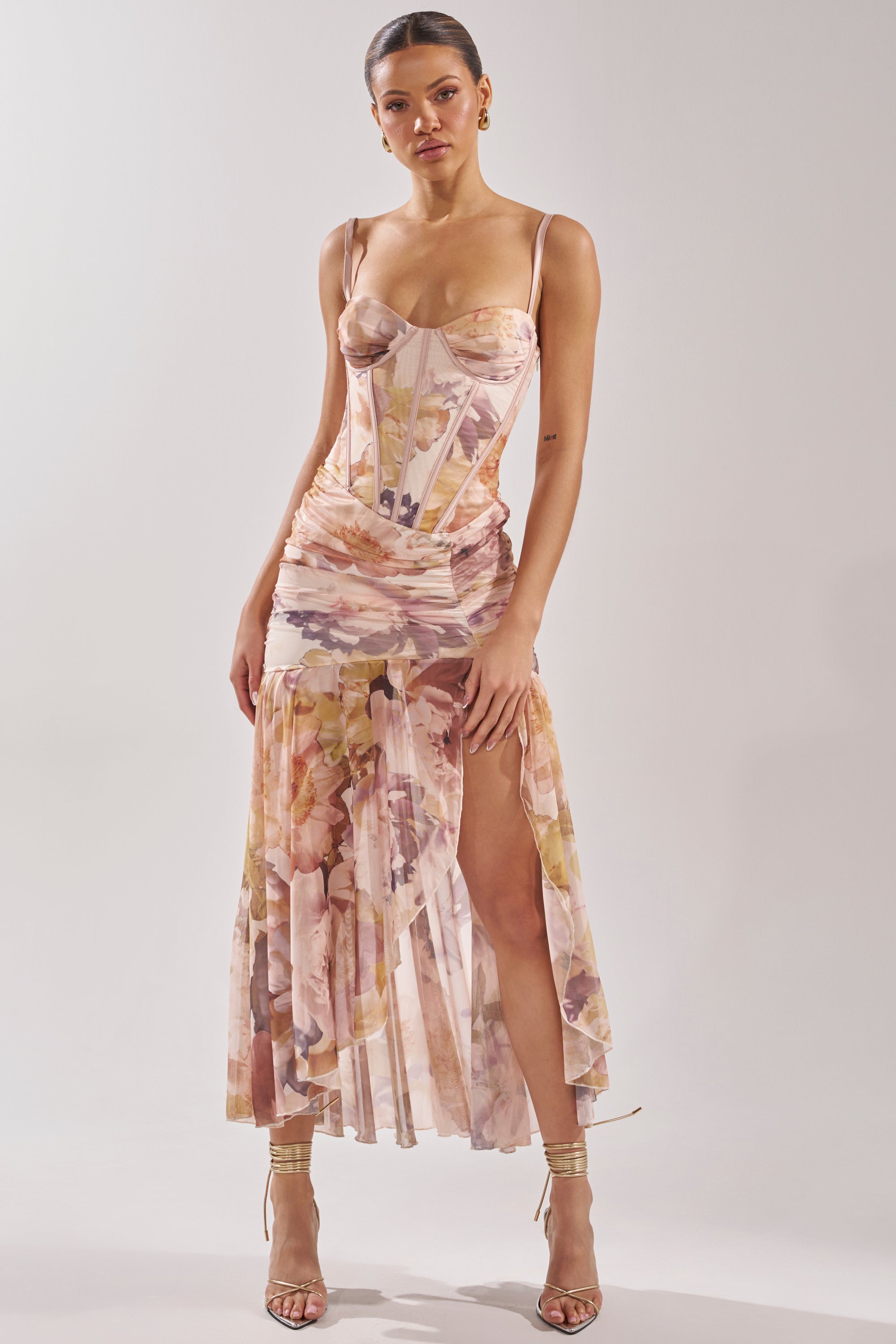 A woman models the WILD FLOWER MAXI DRESS—a sleeveless, watercolor floral dress with a corset bodice and high slit—paired with strappy heels against a plain light background. 1