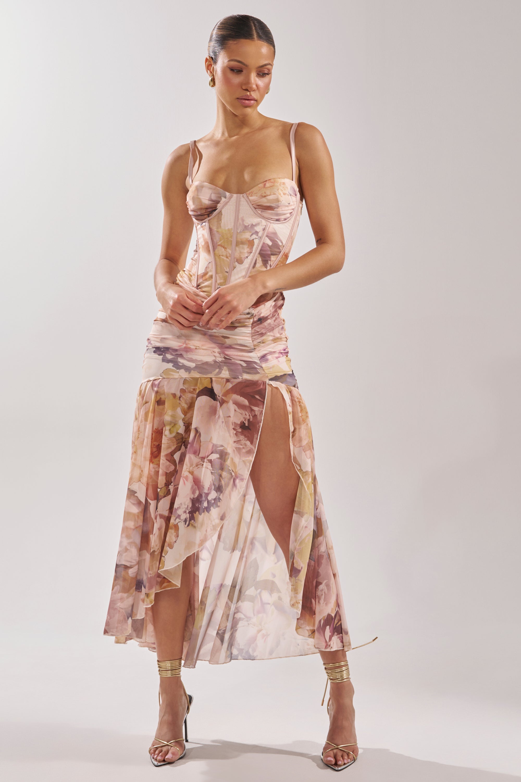 A woman stands in strappy gold heels against a plain light background, wearing the WILD FLOWER MAXI DRESS—a fitted mesh maxi with a corset bodice and watercolor floral print. 3