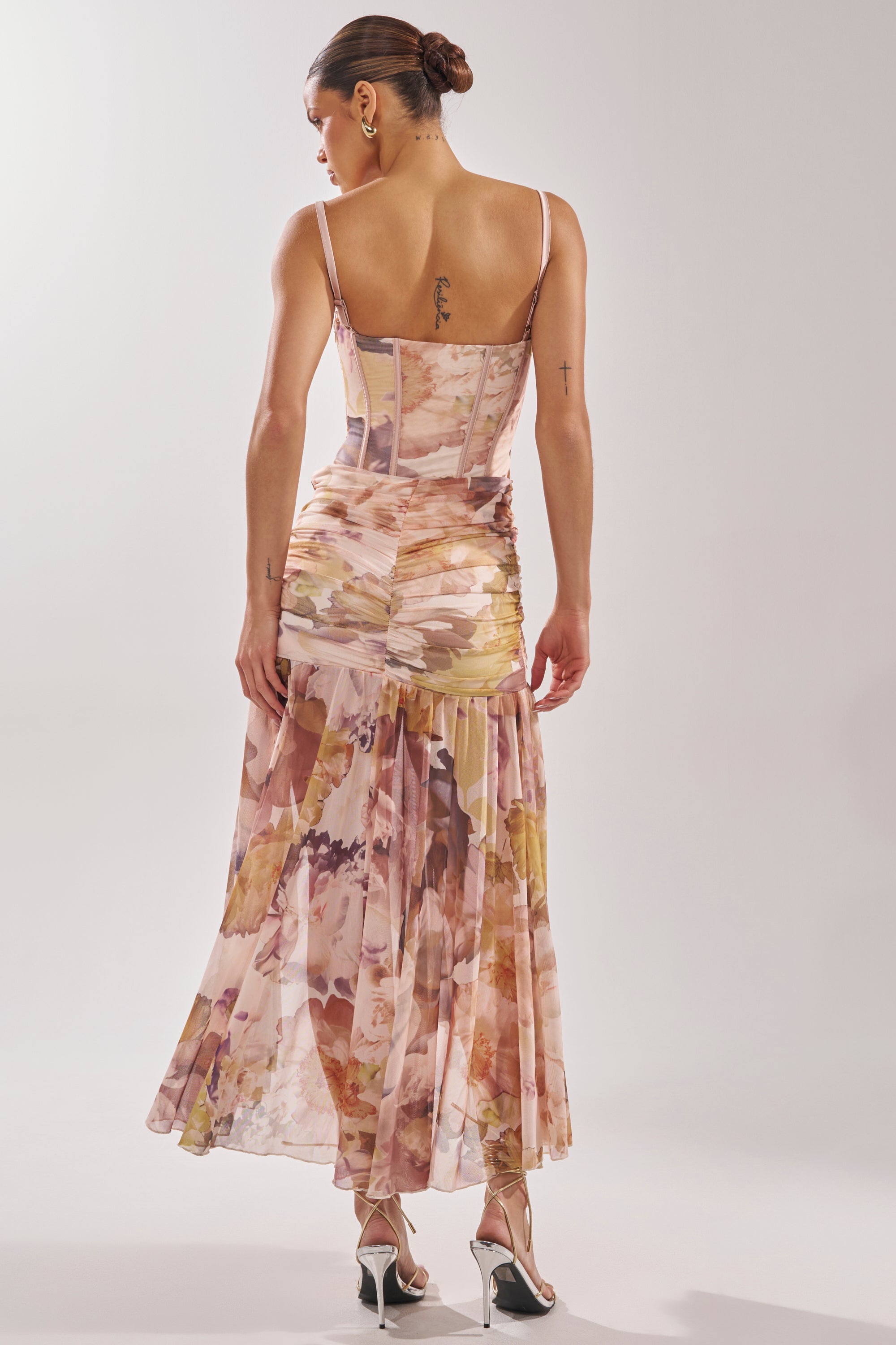 A woman wears the WILD FLOWER MAXI DRESS, a fitted watercolor floral dress with spaghetti straps, revealing her back tattoos. She styles this elegant wedding guest look with heeled sandals and a neat bun. 5