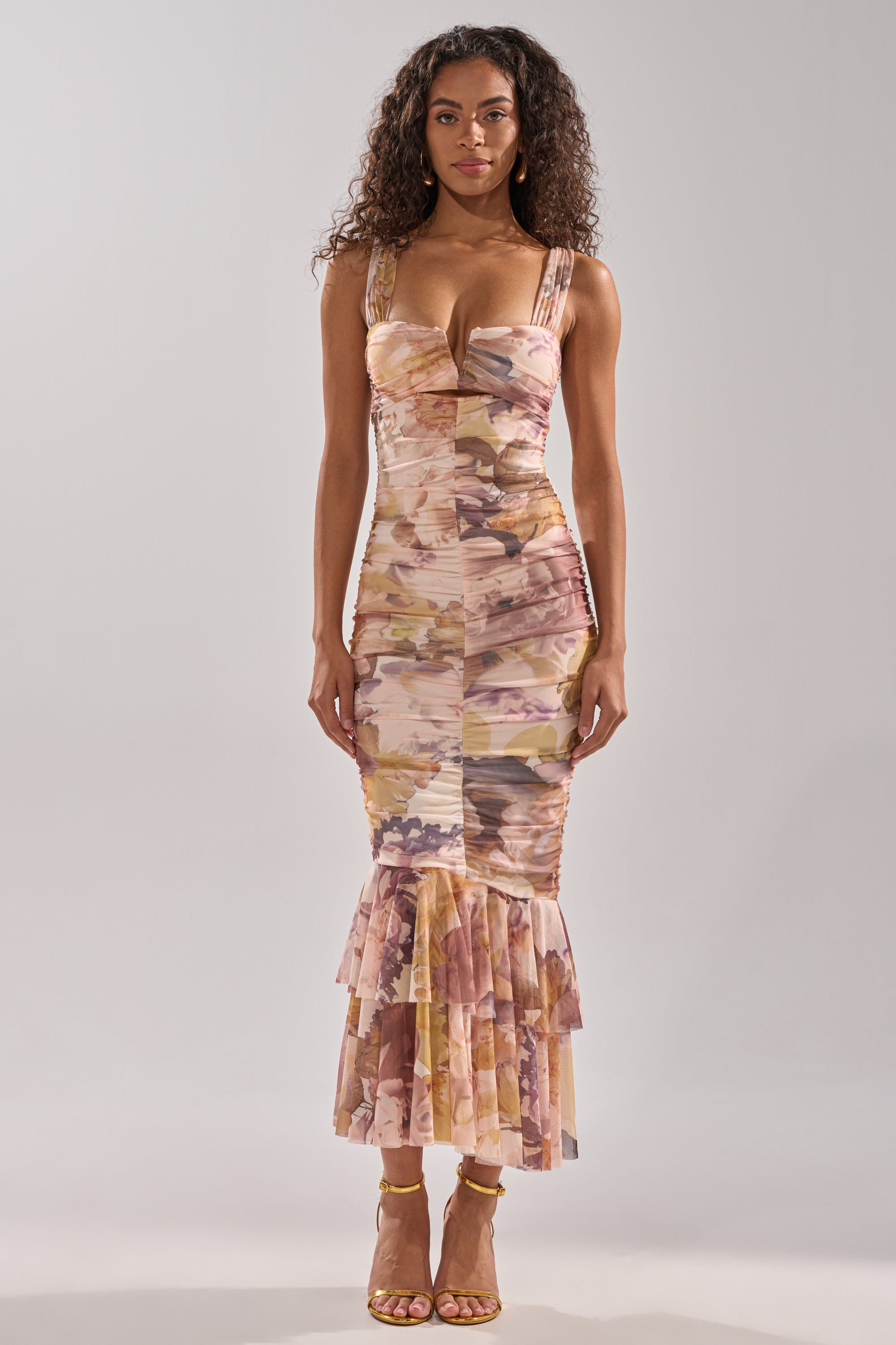 A woman with long curly hair and a neutral expression faces forward in the LOVER GIRL SLINKY MIDI DRESS, showcasing a pastel floral print, ruffled hem, and yellow strappy heels against a plain background.