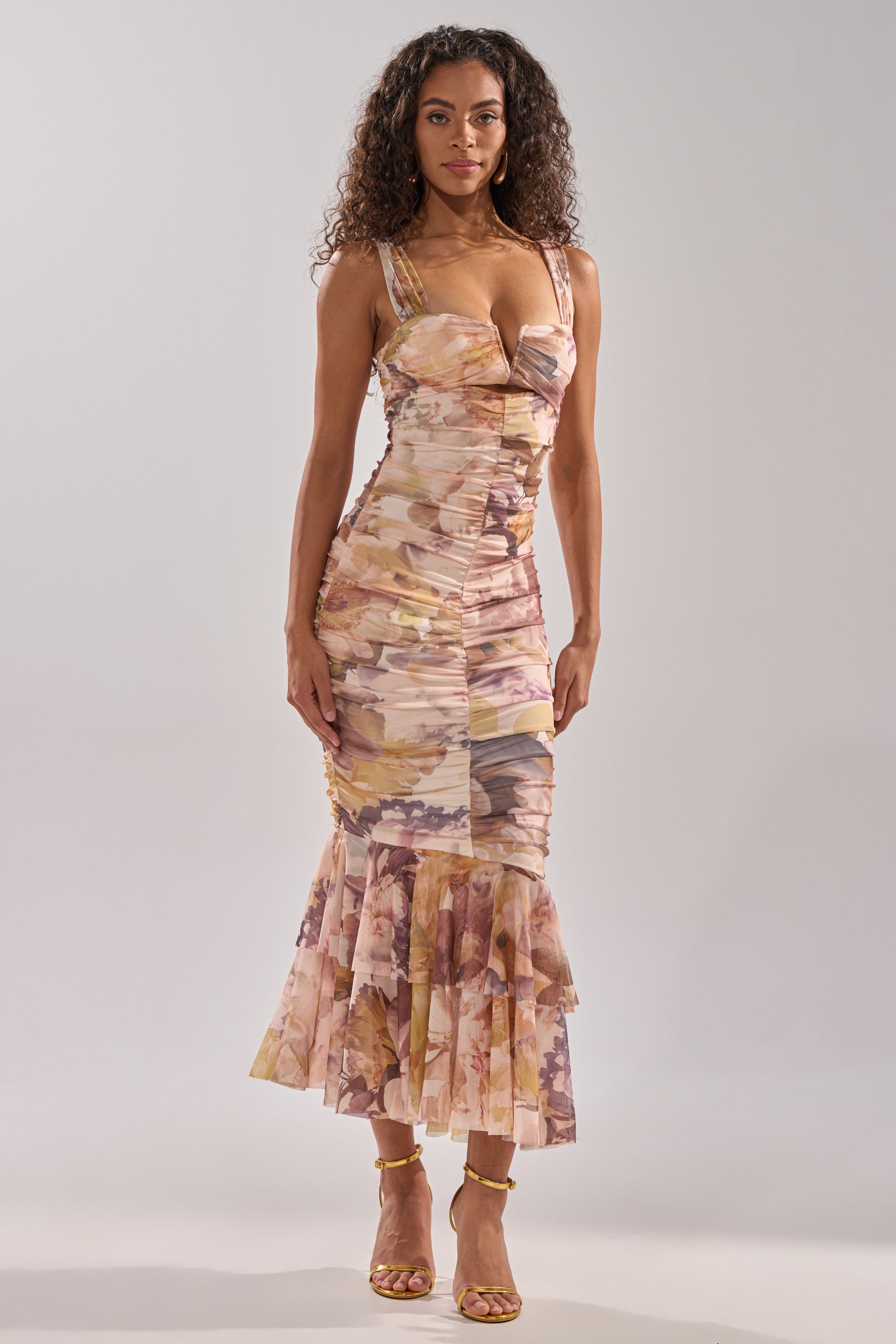 A woman with curly hair confidently models the LOVER GIRL SLINKY MIDI DRESS—a fitted, sleeveless floral dress with a ruffled mermaid hem—paired with gold strappy heels against a plain light background.
