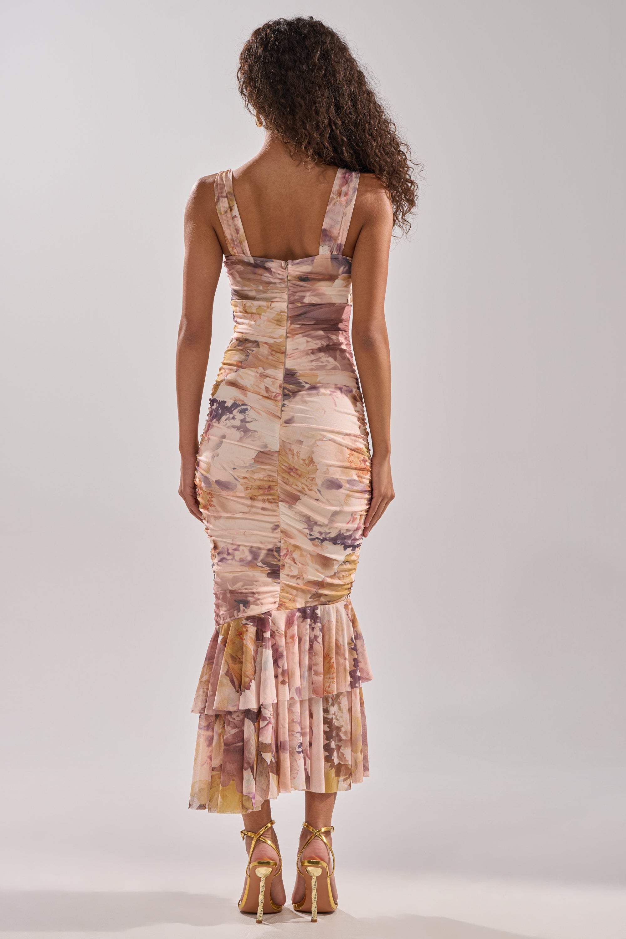 A woman with curly hair stands with her back to the camera in strappy heels, wearing the LOVER GIRL SLINKY MIDI DRESS featuring a floral print and ruffled hem.