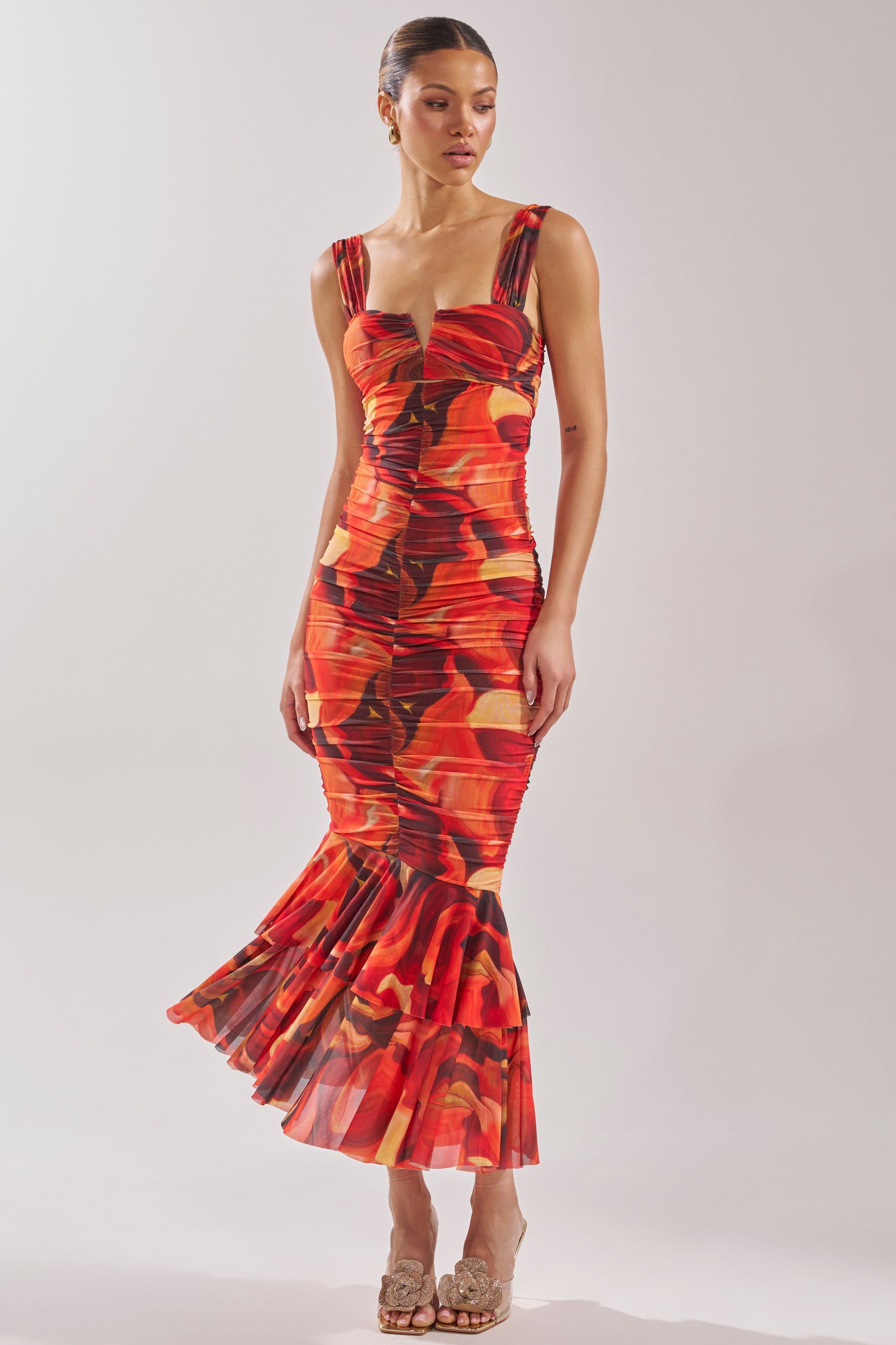 A woman in the BELONG IN A MUSEUM RED MAXI DRESS, featuring a form-fitting silhouette, sleeveless design, flame print, and flared hem, stands against a light background with open-toe heels. 1