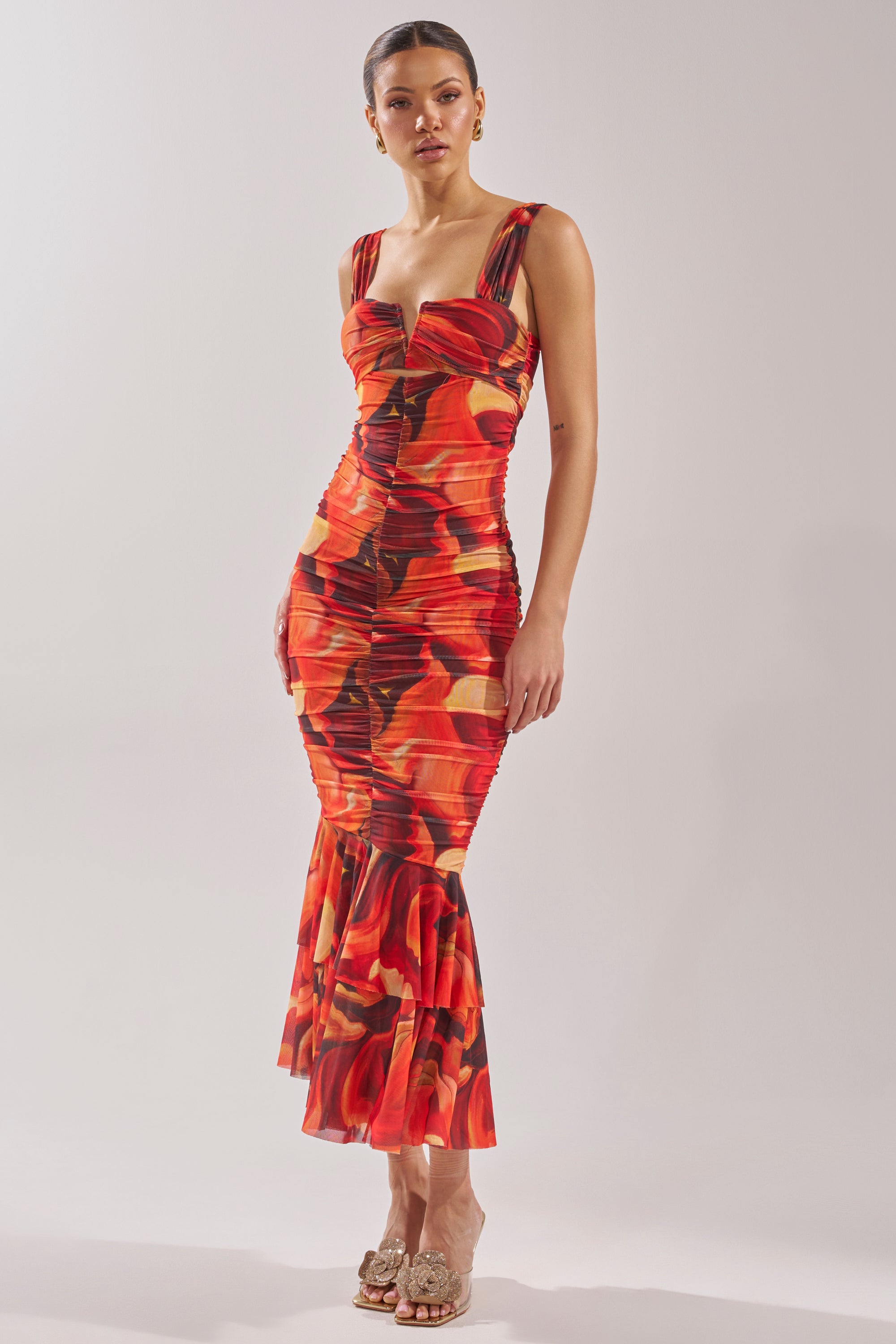 A woman poses against a plain light background in the BELONG IN A MUSEUM RED MAXI DRESS—fitted, sleeveless, with a flared hem. Her hair is pulled back and she accessorizes with gold earrings and heeled sandals. 2