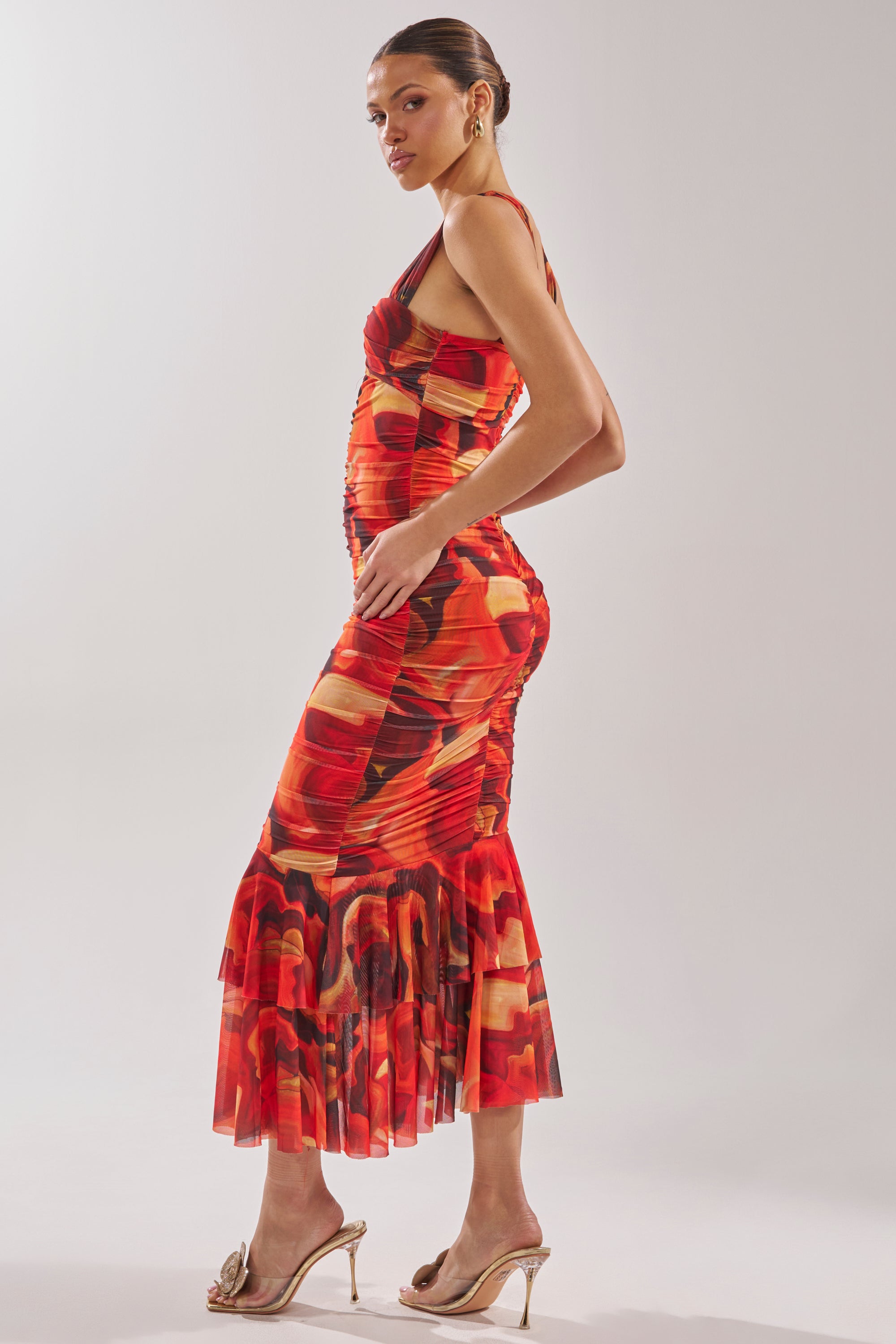 A woman stands sideways in the BELONG IN A MUSEUM RED MAXI DRESS, a fitted sleeveless style with a ruffled hem, paired with gold high-heeled sandals. Her hair is styled in a sleek bun and she wears hoop earrings. 4