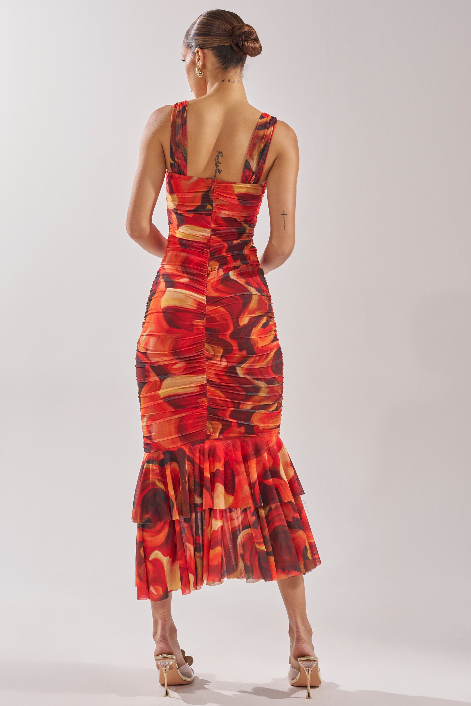 A woman with her hair in a bun models the BELONG IN A MUSEUM RED MAXI DRESS, featuring a form-fitting flame print and ruffled hem, shown from the back with beige high-heeled sandals. 5