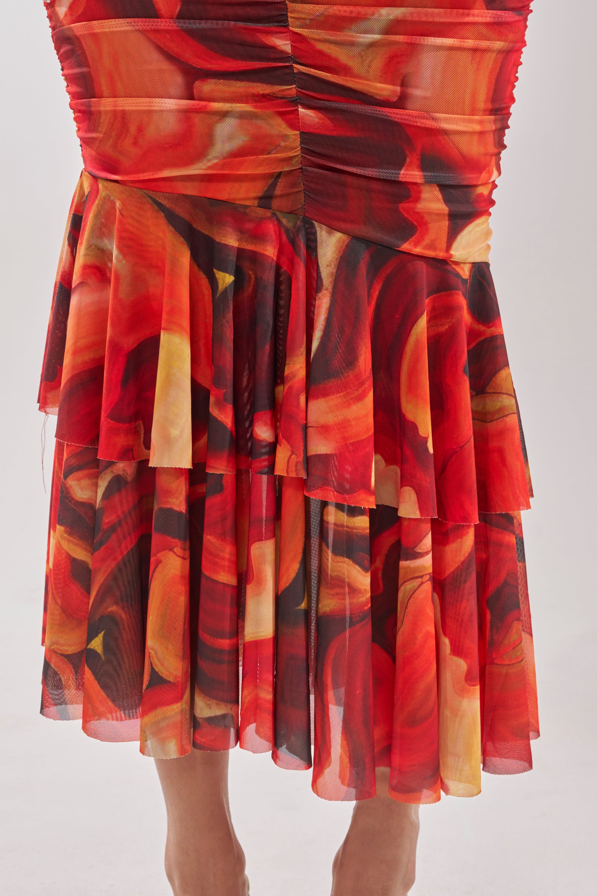 A close-up of the BELONG IN A MUSEUM RED MAXI DRESS shows vibrant red, orange, yellow, and black abstract patterns. The gathered, pleated fabric mimics fiery flames for a dynamic, textured look. 7