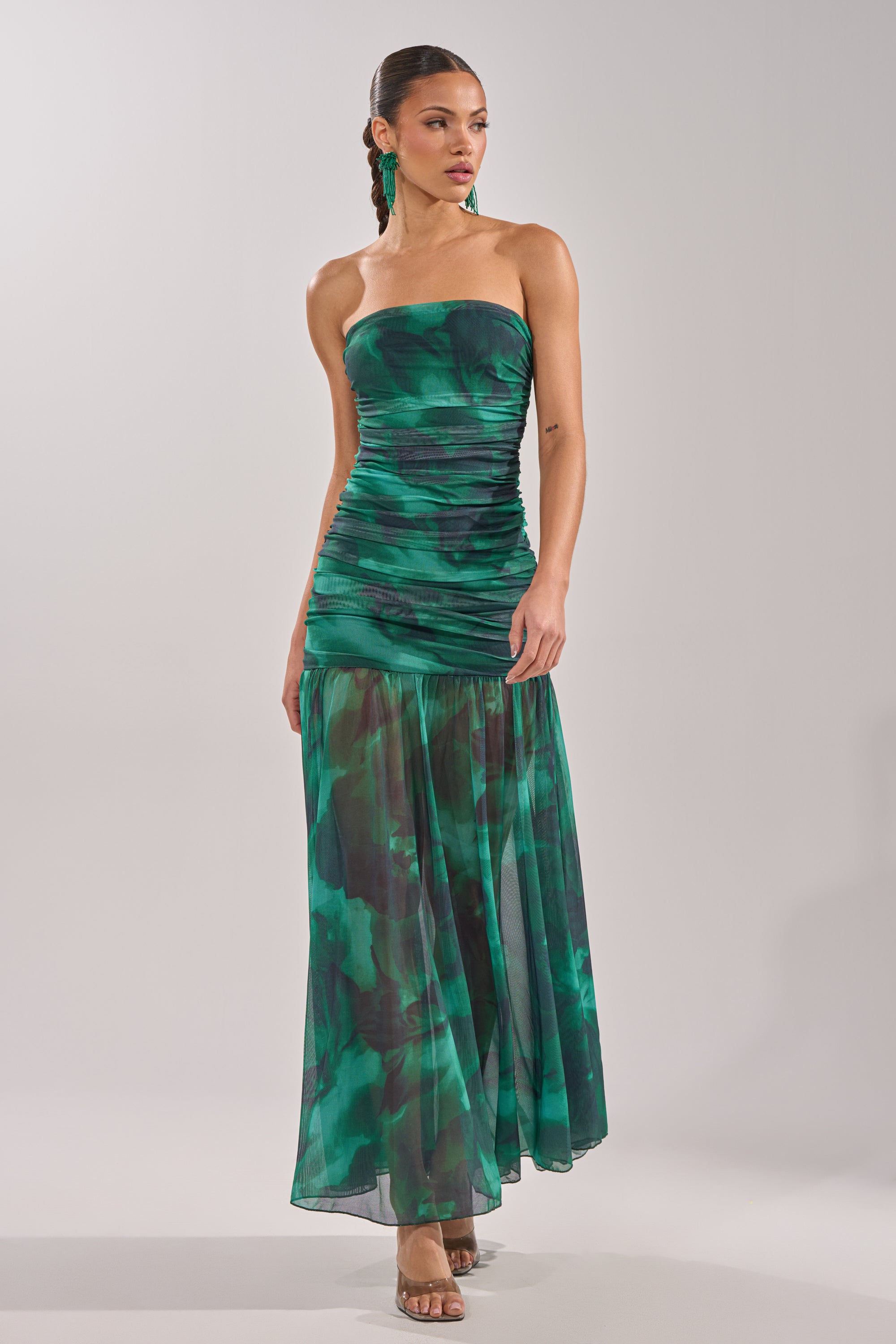 A woman wears the UNDER THE SEA MAXI DRESS—a strapless mesh gown with a ruched bodice, green and black patterns, and a sheer flowing skirt. Her hair is pulled back, she accessorizes with green earrings and neutral heels against a light backdrop. 2