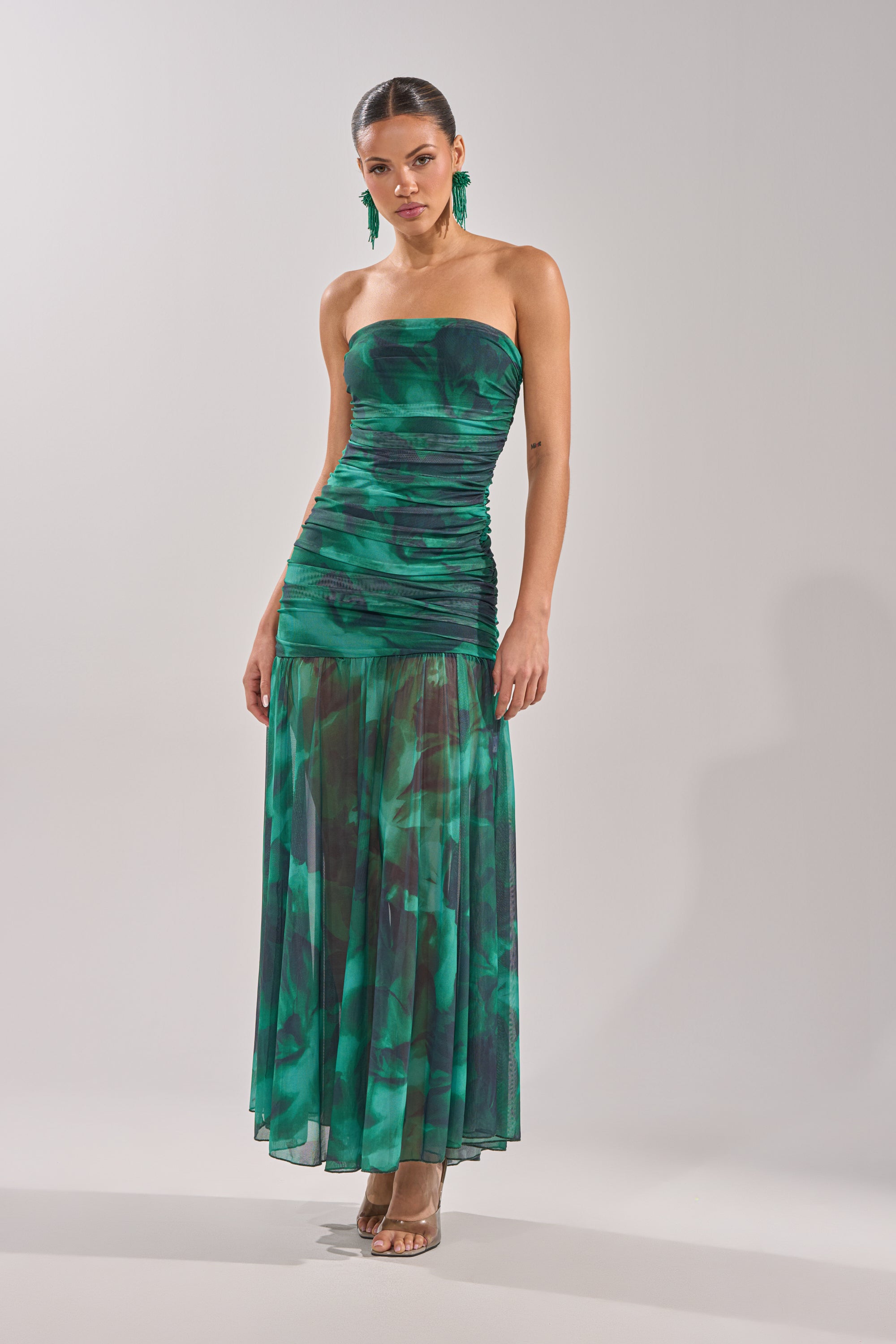 A woman in the UNDER THE SEA MAXI DRESS, featuring a strapless ruched bodice and sheer maxi skirt, poses against a plain backdrop with her hair pulled back, accessorized with green earrings and clear-heeled shoes. 3