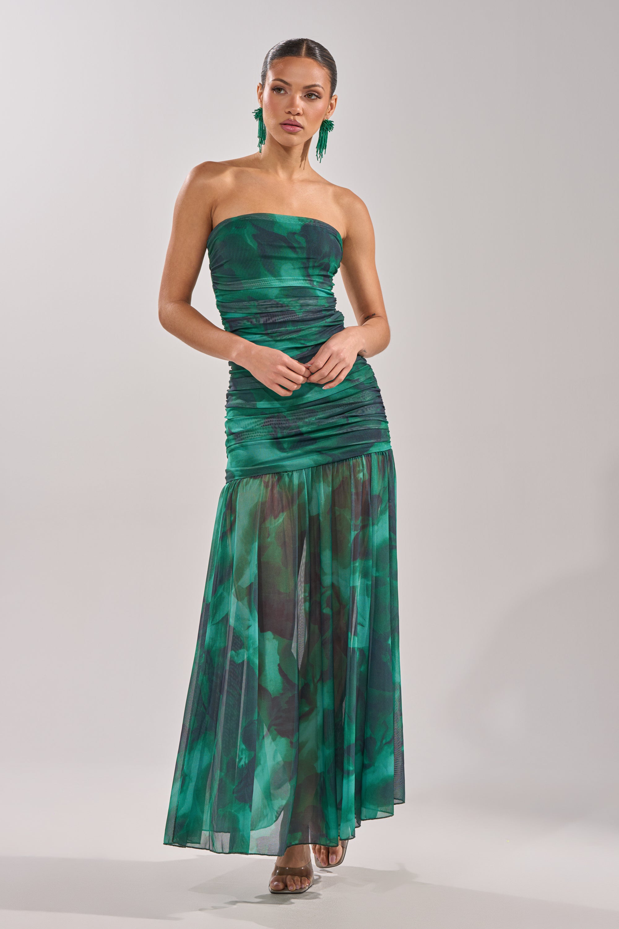 A woman faces forward wearing the UNDER THE SEA MAXI DRESS—strapless, with a ruched bodice and sheer, flowing skirt—paired with green statement earrings and heeled sandals. 4