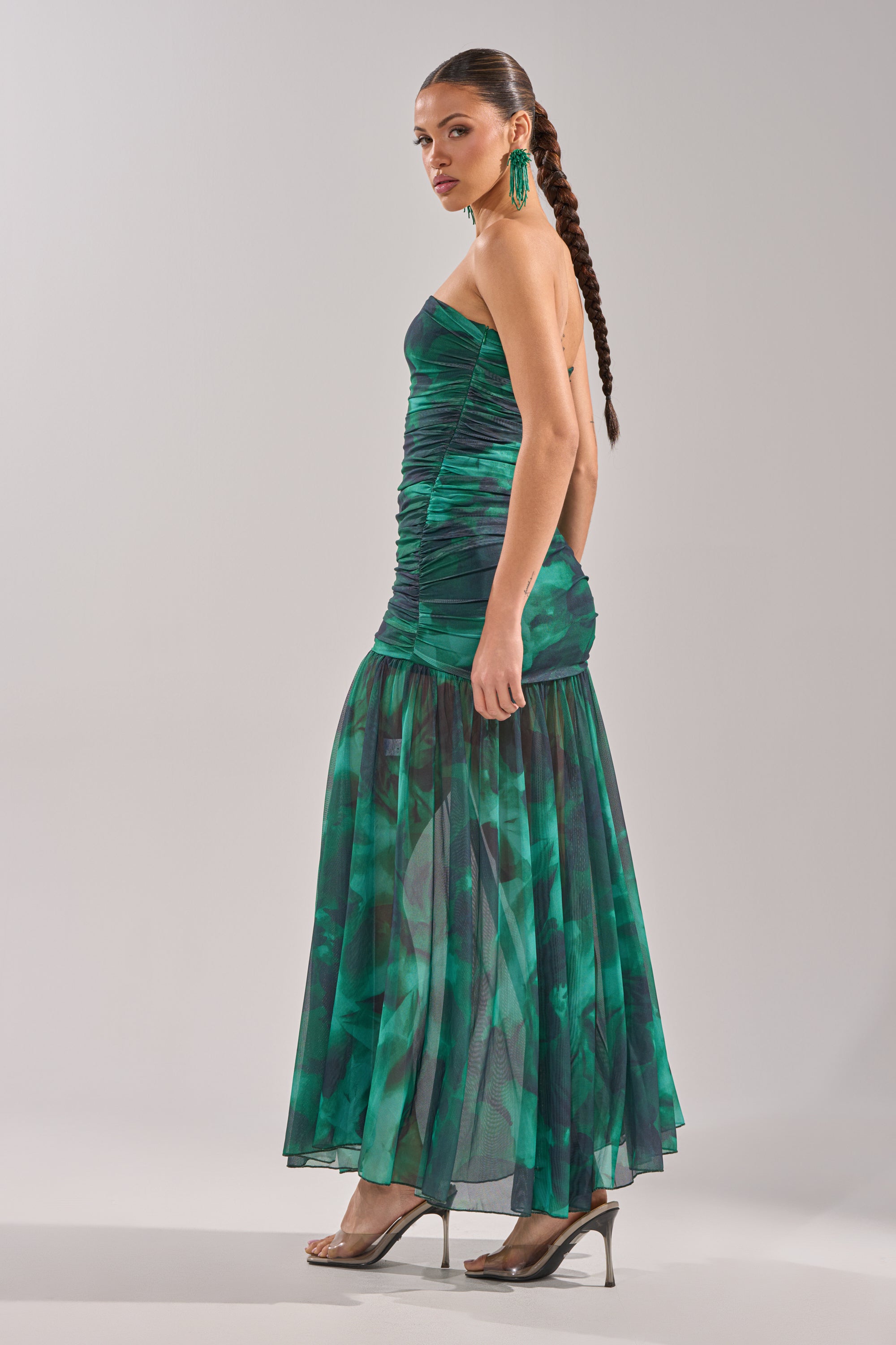 A woman with a long braided ponytail models the UNDER THE SEA MAXI DRESS, showcasing its ruched bodice and strapless, green and blue patterned silhouette, paired with statement earrings and clear heels against a light background. 5