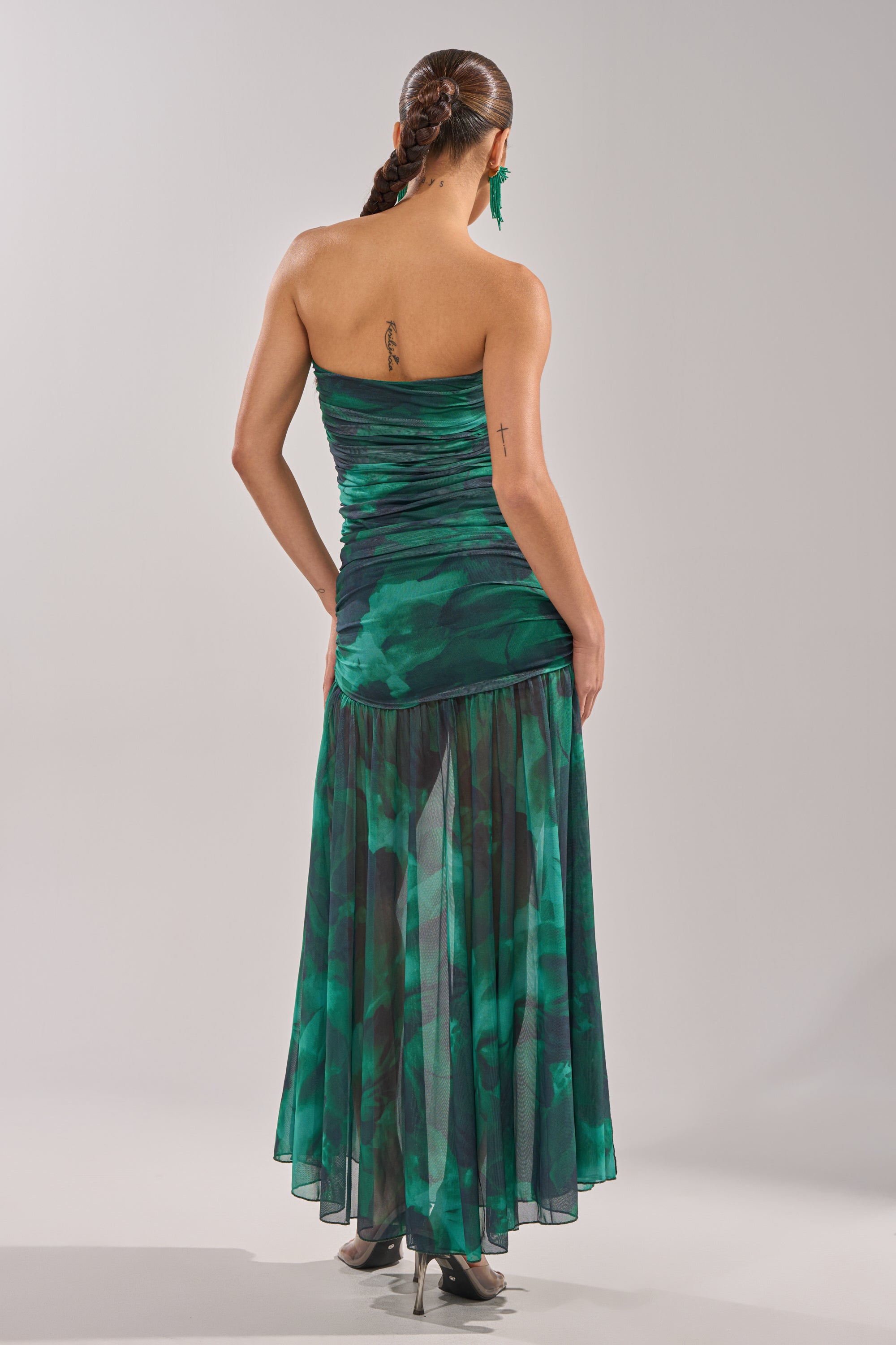 A woman with braided hair stands facing away, wearing the UNDER THE SEA MAXI DRESS with a flowing sheer skirt and high heels. Visible tattoos are on her upper back and left arm, set against a plain, light-colored background. 7