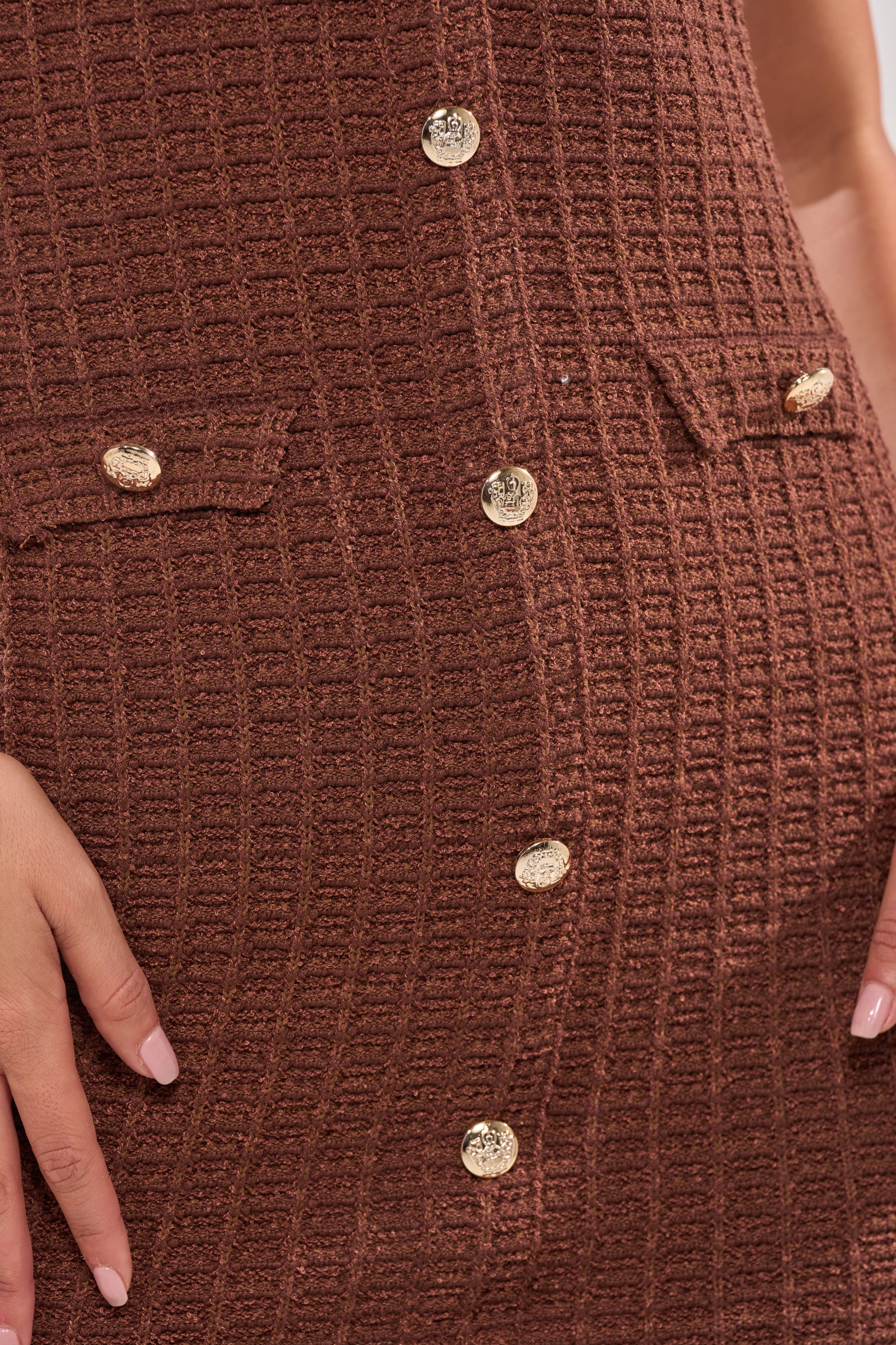 Close-up of the BUTTONED DOWN BADDIE MINI DRESS IN BROWN, featuring a brown waffle knit, gold center buttons, and two small flap pockets. A person’s hands rest at their sides, partially visible in the frame. 6