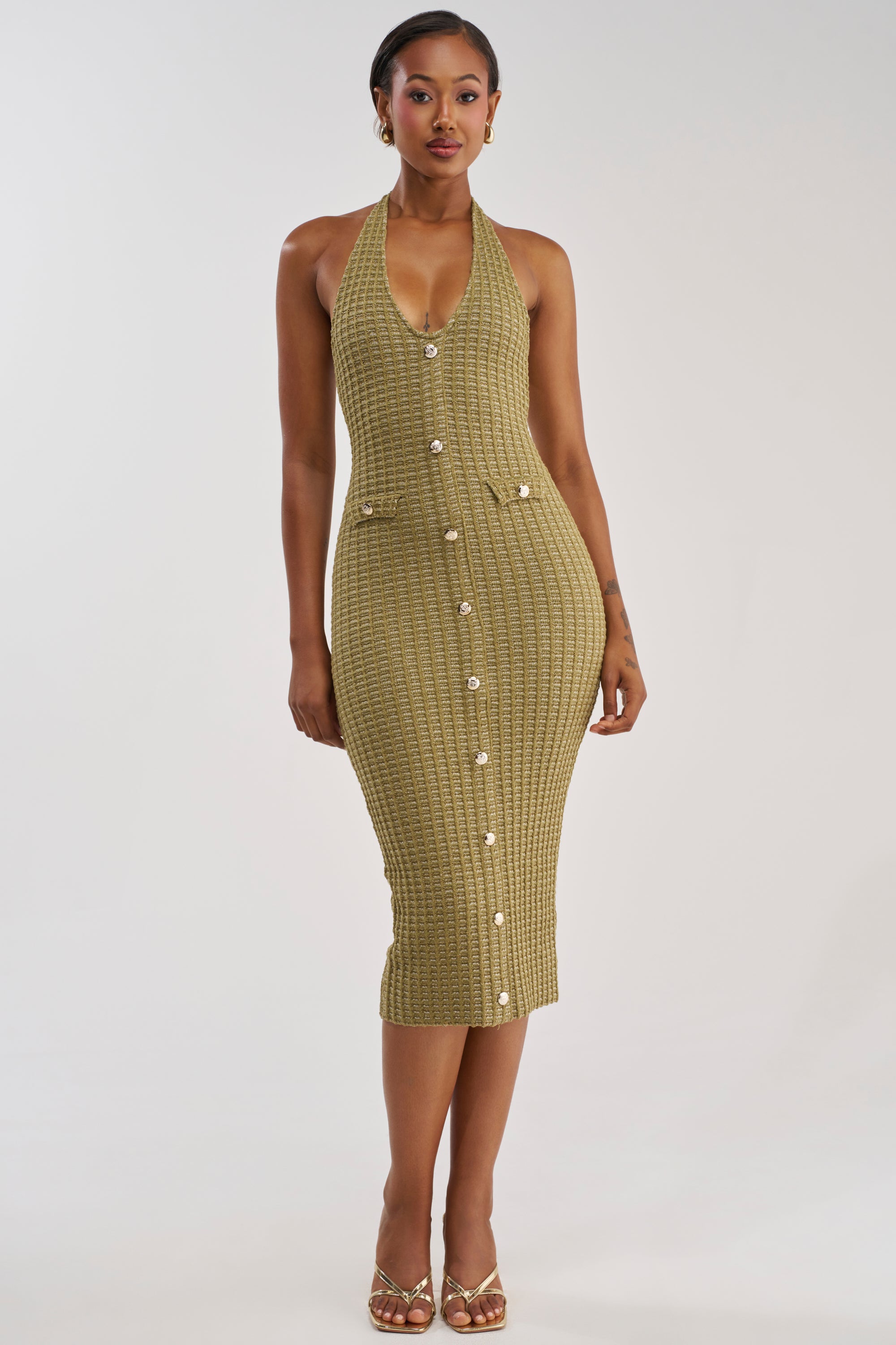 Model wearing the BUTTONED DOWN BADDIE MIDI DRESS IN OLIVE—a textured, halter-neck, button-down midi dress—styled with gold earrings and strappy heels, posed against a plain white background.