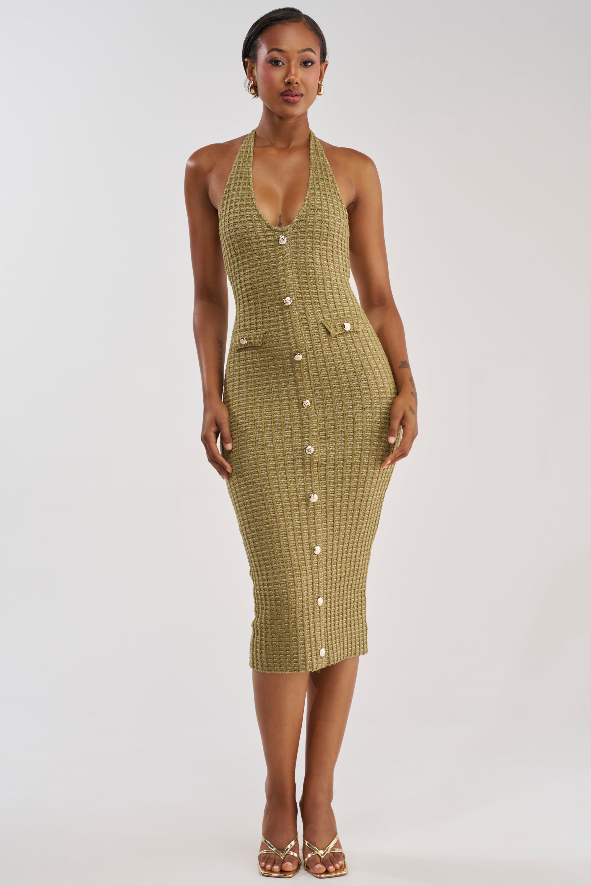 A woman stands facing forward against a plain white background, modeling the BUTTONED DOWN BADDIE MIDI DRESS IN OLIVE. The sleeveless, olive green halter midi dress features buttons down the front and two pockets, creating a bodycon silhouette that flatters her figure. She wears gold earrings and gold sandals, with short hair completing her look.