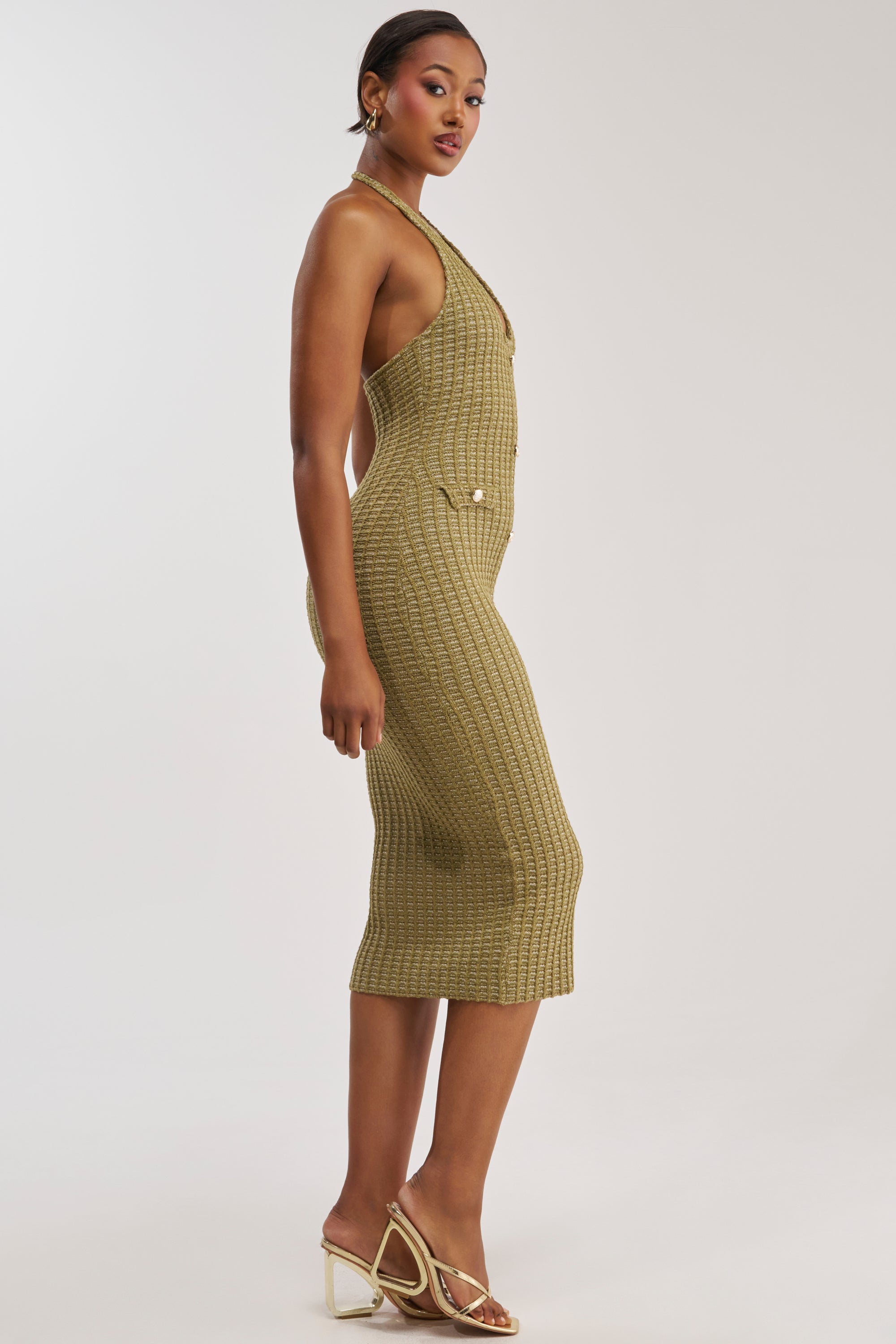 Model poses in profile wearing the BUTTONED DOWN BADDIE MIDI DRESS IN OLIVE, a fitted sleeveless knit dress styled with gold strappy heels and accessorized with gold earrings. She has short hair and stands against a plain light background.