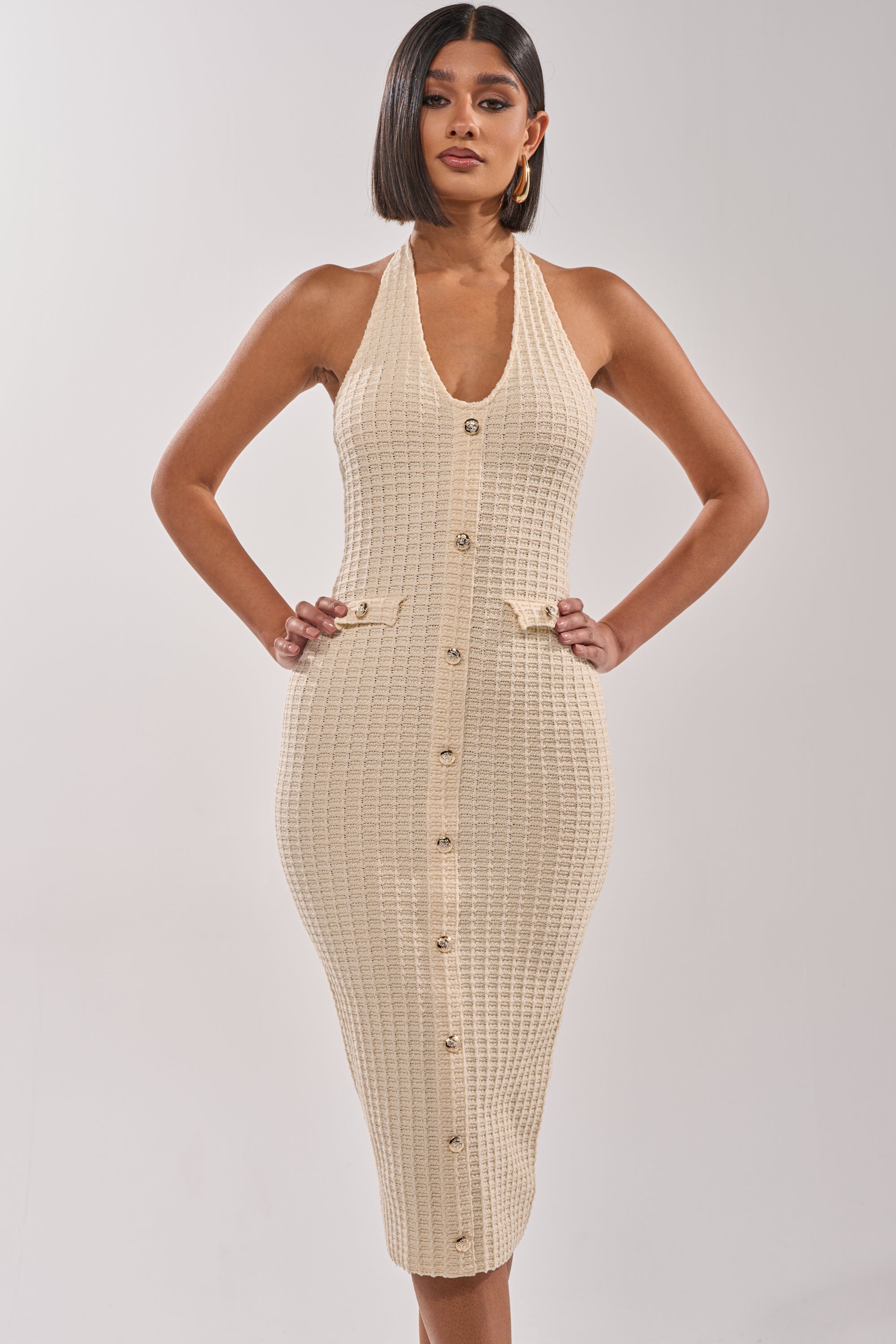 A woman models the BUTTONED DOWN BADDIE MIDI DRESS IN WHITE, a fitted sleeveless knit summer party dress with a V-neckline, gold button details, and decorative waist bows. She has straight dark shoulder-length hair and wears hoop earrings. 1