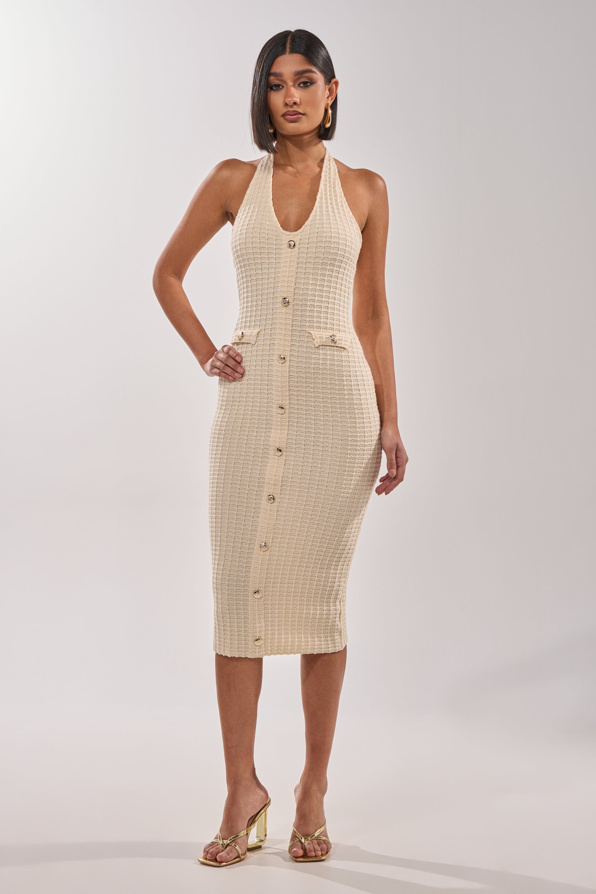 A woman faces forward in the BUTTONED DOWN BADDIE MIDI DRESS IN WHITE—a sleeveless knit dress with gold buttons and two small front pockets. She has straight dark hair, gold heels, and hoop earrings, posing against a plain background. 4