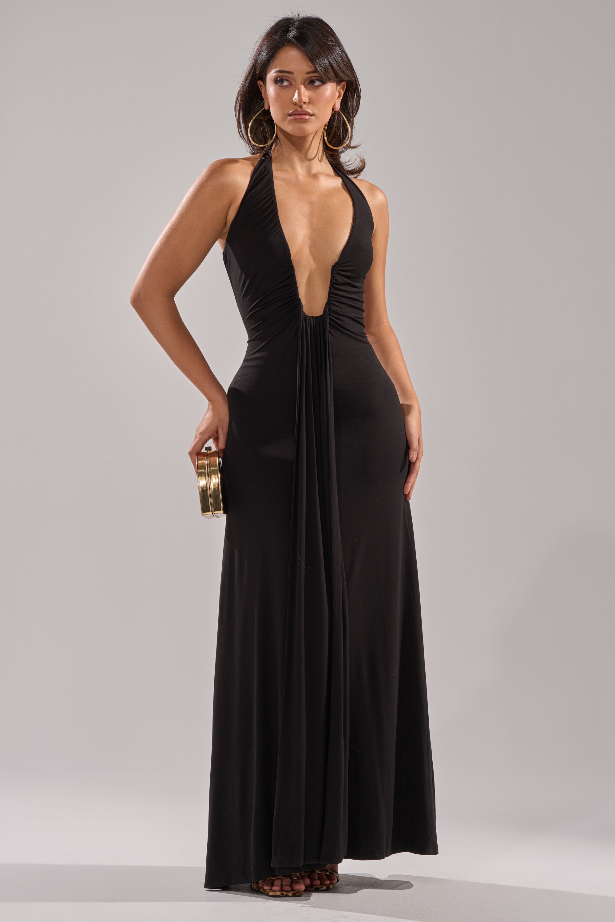 A woman models the MAKE YOU LOSE YOUR MIND MAXI DRESS IN BLACK, a sleeveless floor-length halter dress with a sheath silhouette and deep plunging neckline, holding a gold clutch against a plain light gray background.