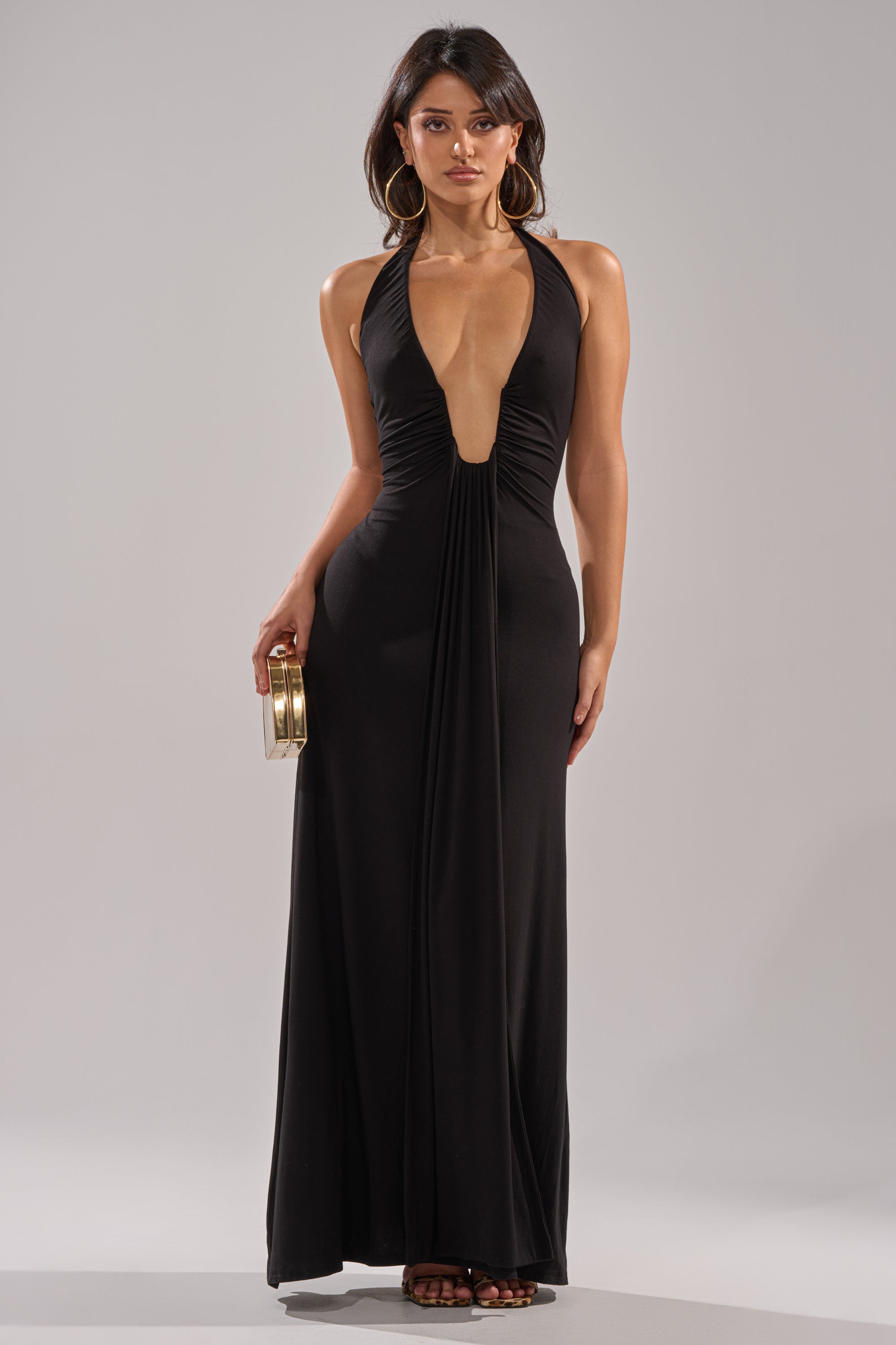 A woman wears the MAKE YOU LOSE YOUR MIND MAXI DRESS IN BLACK, a sleeveless deep V-neck maxi, facing forward with a gold clutch in her right hand, large hoop earrings, and long loose hair.