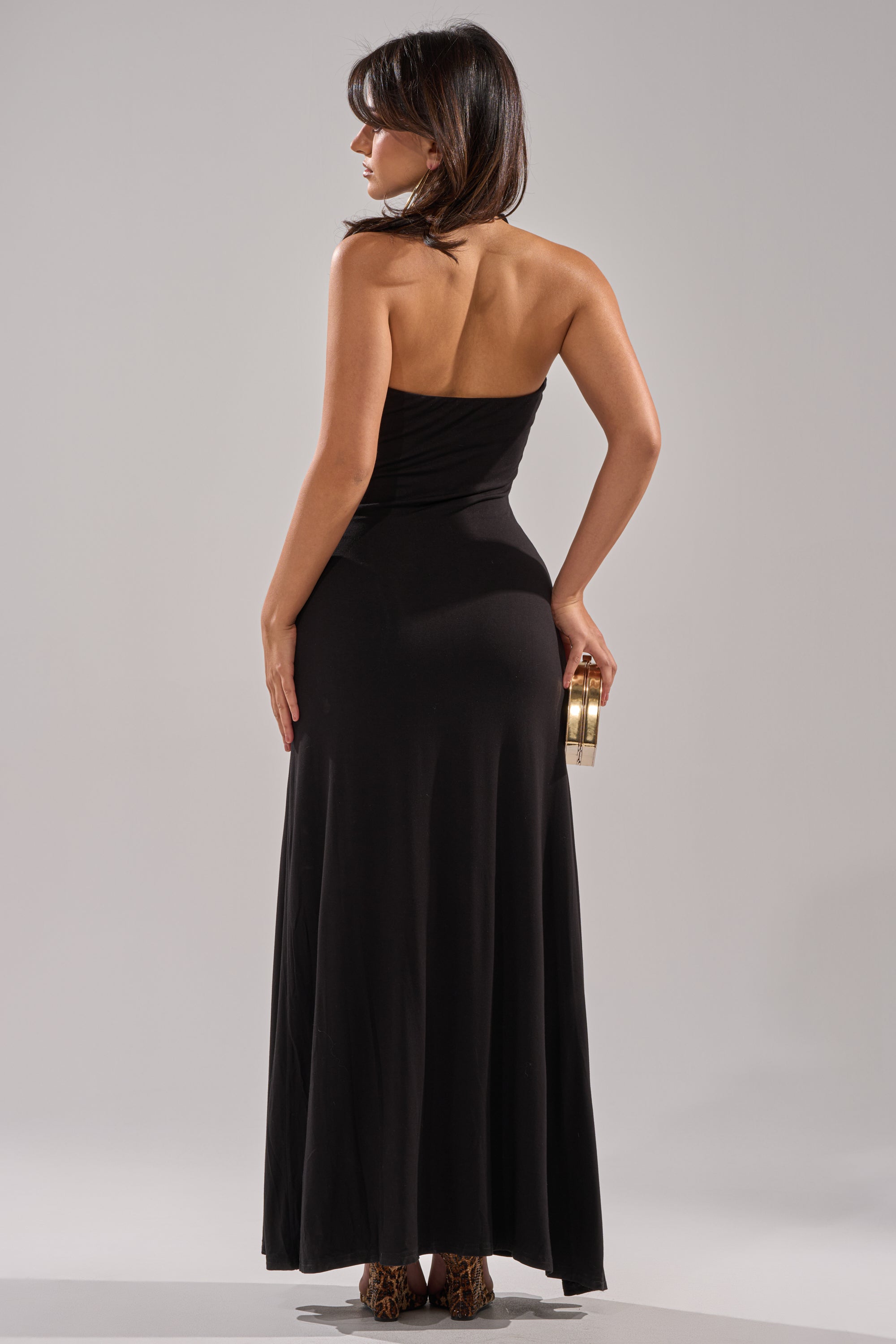 A woman with straight dark hair faces away, wearing the MAKE YOU LOSE YOUR MIND MAXI DRESS IN BLACK, a sheath-style halter maxi dress, and holds a small gold clutch in her right hand against a plain light background.