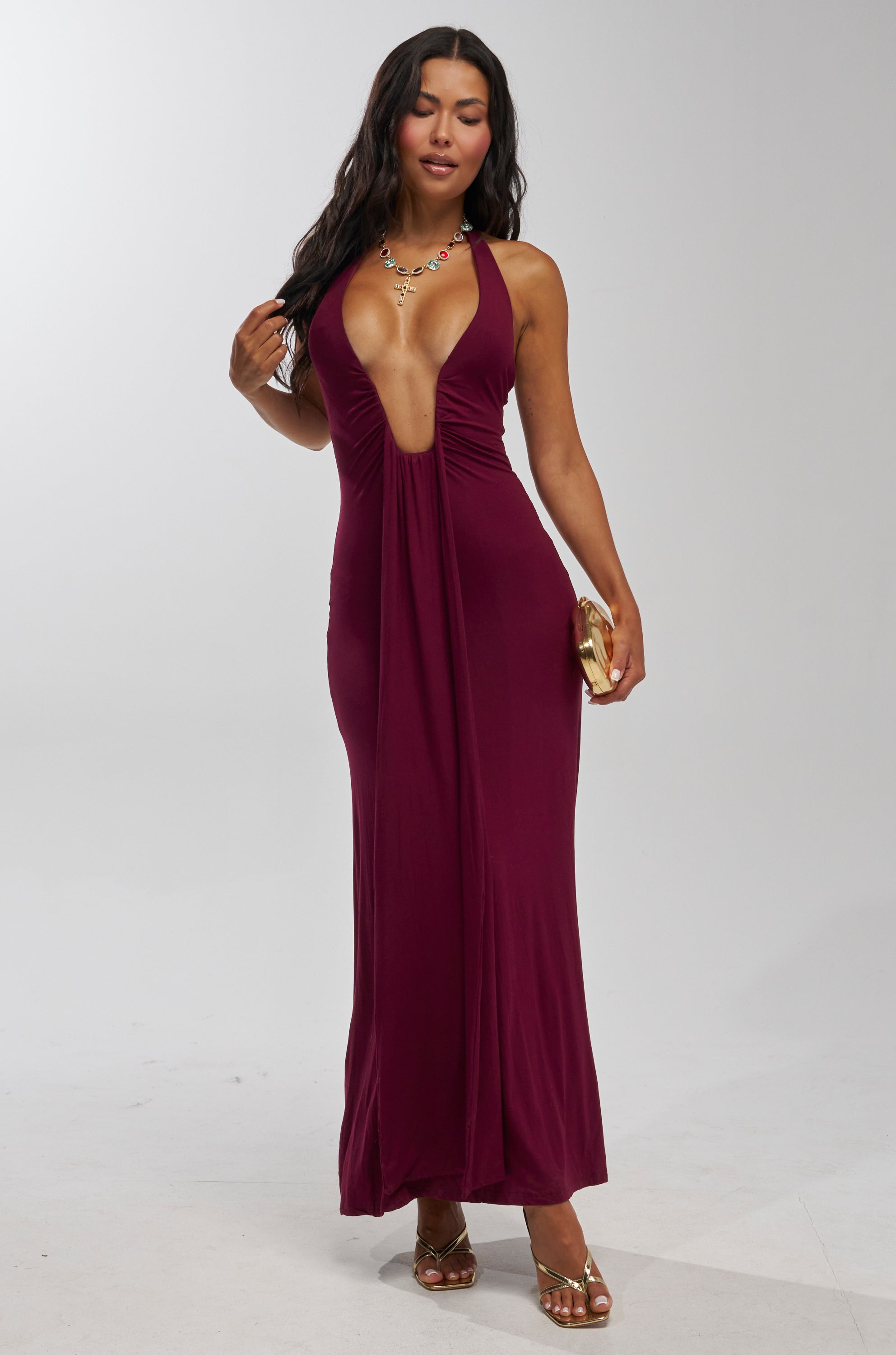 A woman poses in the MAKE YOU LOSE YOUR MIND MAXI DRESS IN BURGUNDY, paired with gold sandals, a statement necklace, and a gold clutch—an elegant vacation evening look against a plain light background.