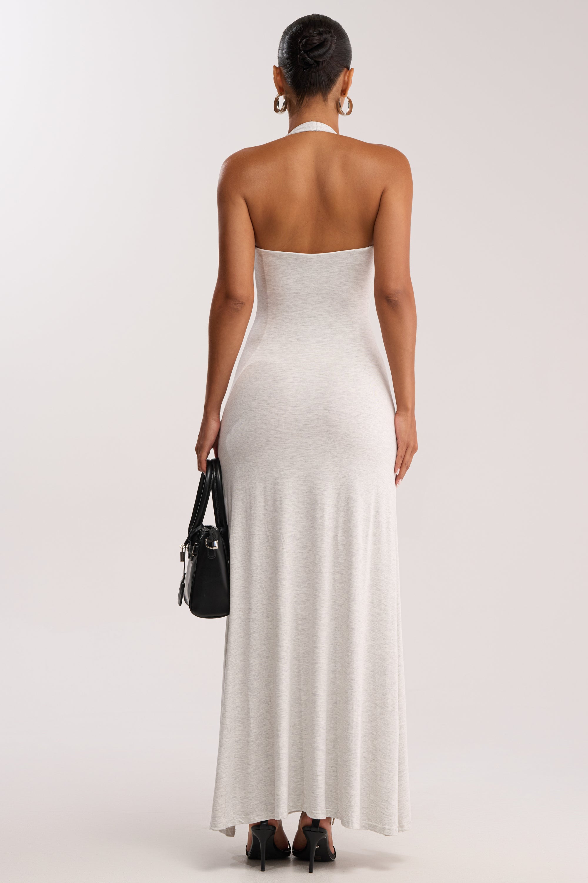 A woman faces away, wearing the MAKE YOU LOSE YOUR MIND MAXI DRESS IN LIGHT GREY—a strapless, floor-length halter dress—styled with black open-toe heels, a black handbag, and her hair in a neat bun.