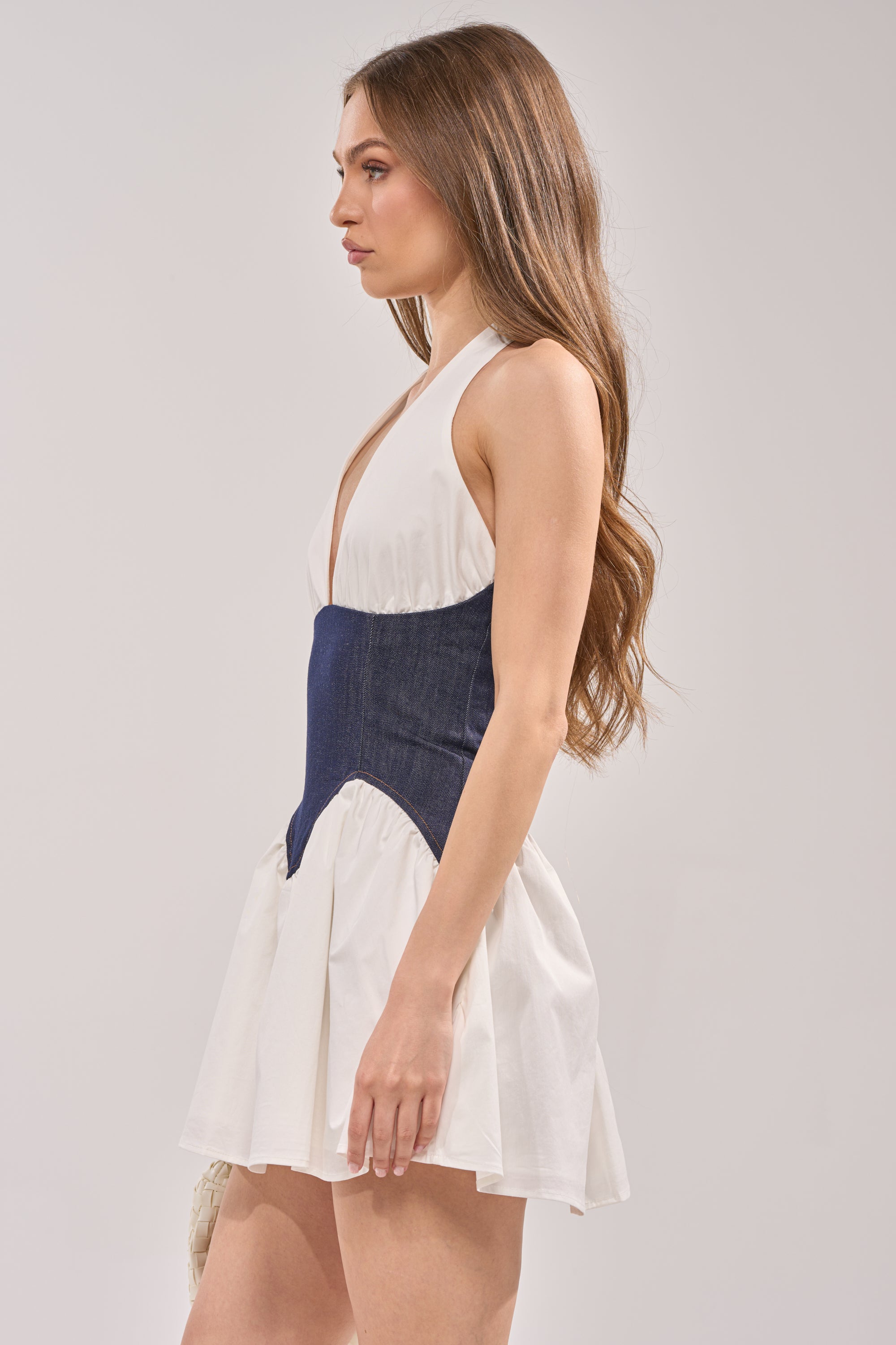 A woman with long brown hair wears the SAY LESS POPLIN MINI DRESS IN WHITE, a sleeveless mini dress with a deep V-neck and fitted dark blue corset-like waist—an ideal choice for spring brunches.