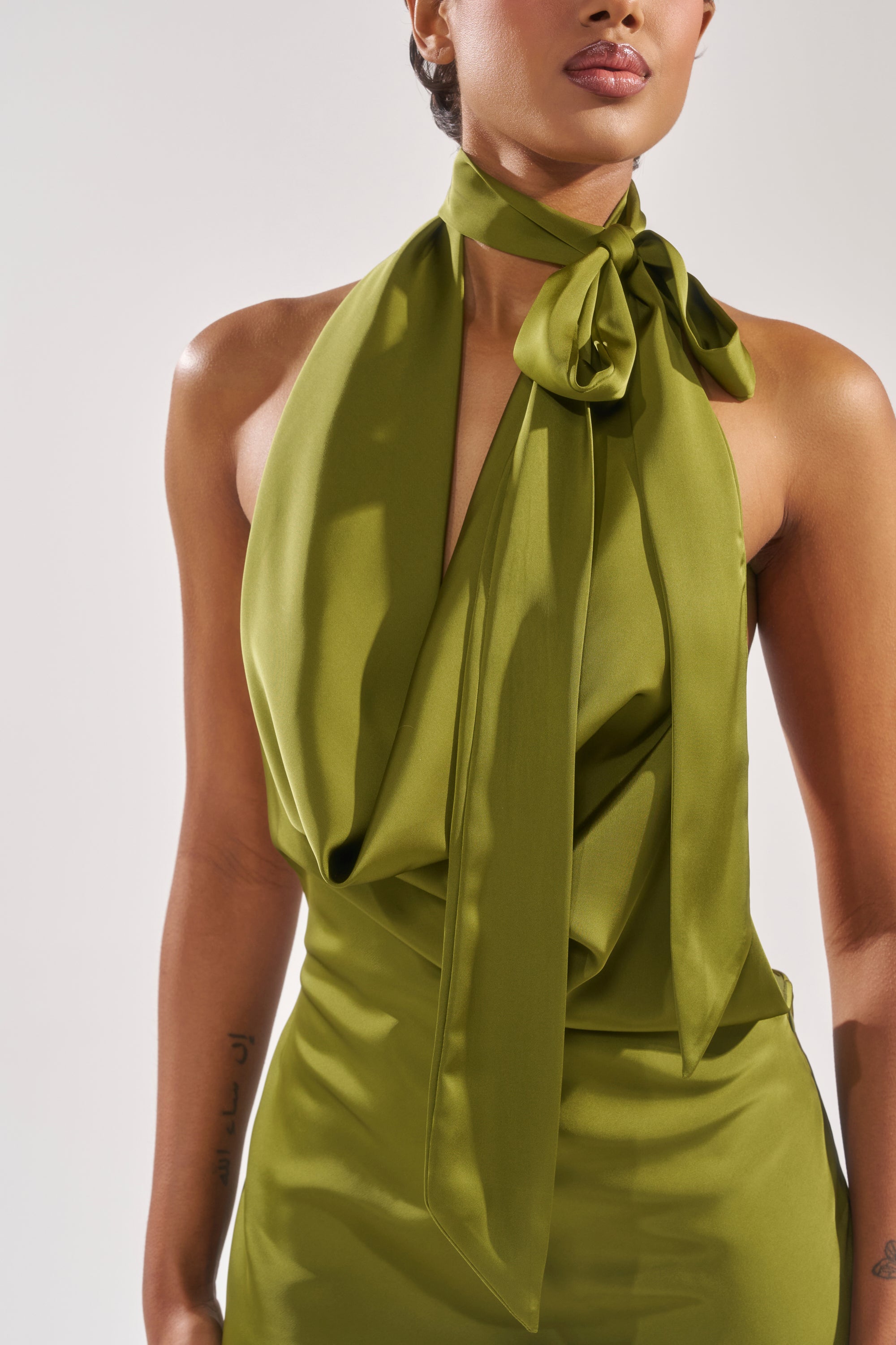 A person wearing the GIGI MINI DRESS IN GREEN—a sleeveless satin mini dress with a draped halter neckline and a large tied bow. Only the upper body is visible against a plain background.