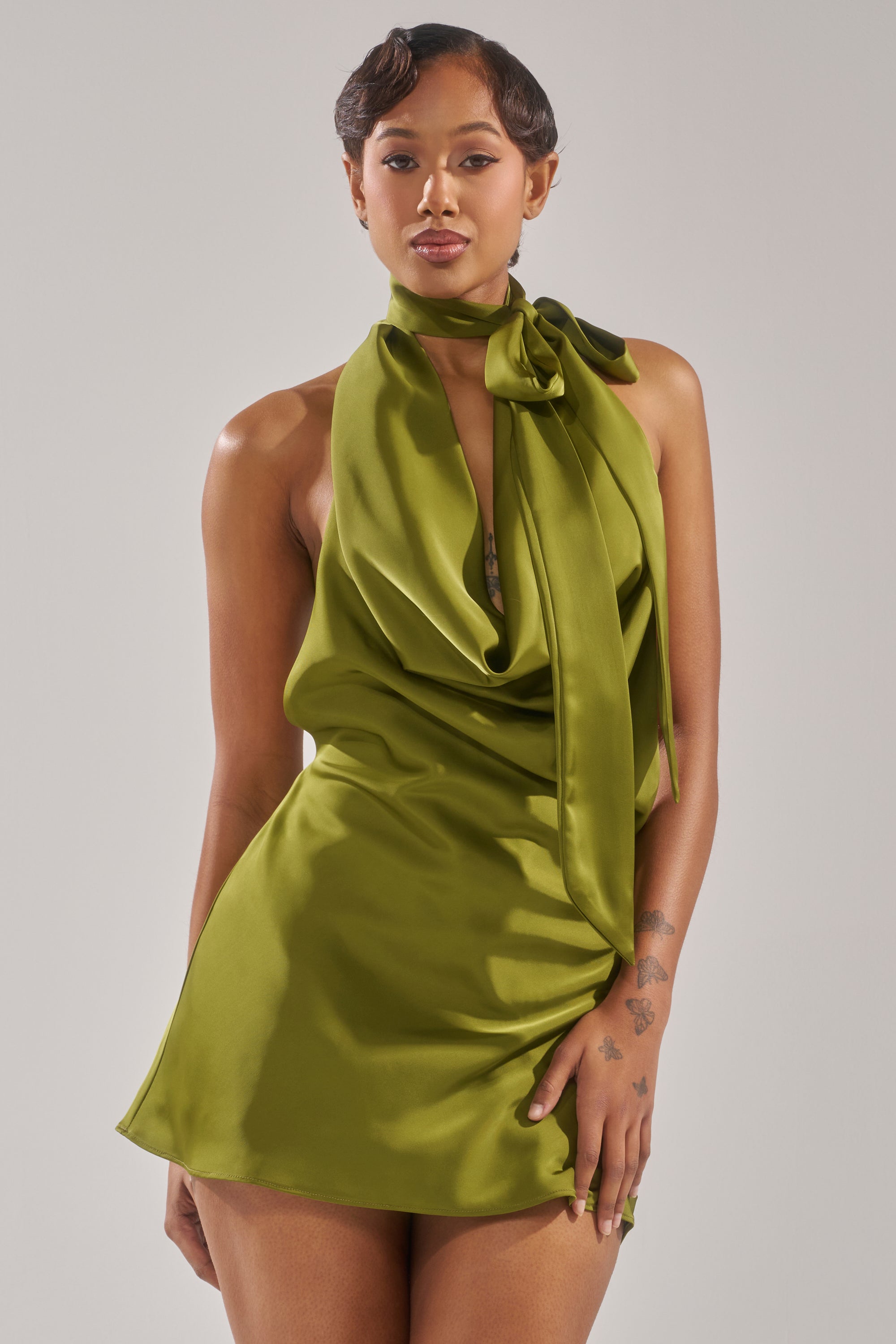 A woman with short hair and visible tattoos wears the GIGI MINI DRESS IN GREEN, a sleeveless satin mini dress featuring a halter cowl neckline, draped front, and large neck bow.