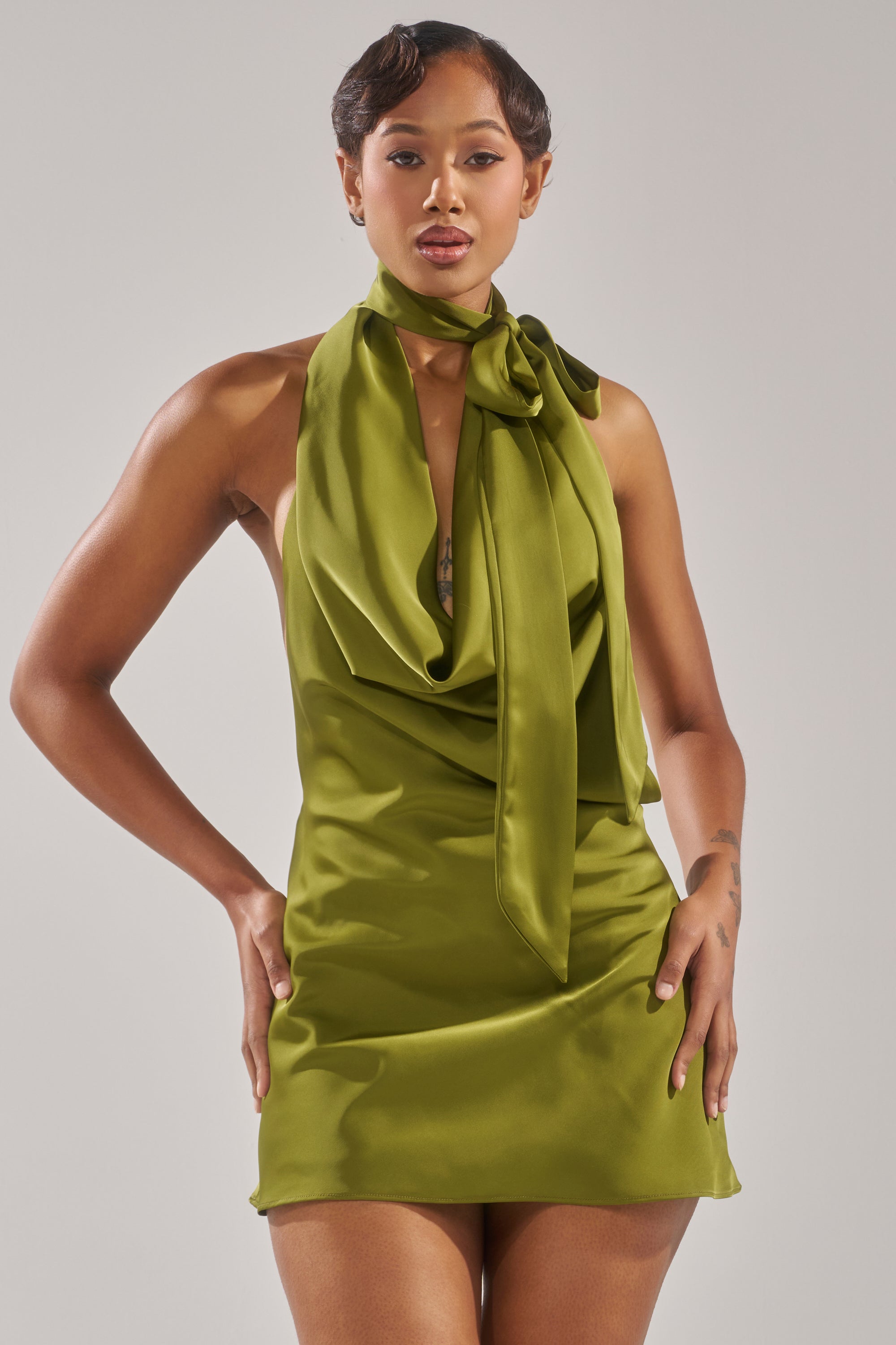 A woman poses confidently in the GIGI MINI DRESS IN GREEN, styled with a halter cowl neckline and an oversized bow at the neck, set against a plain light background.