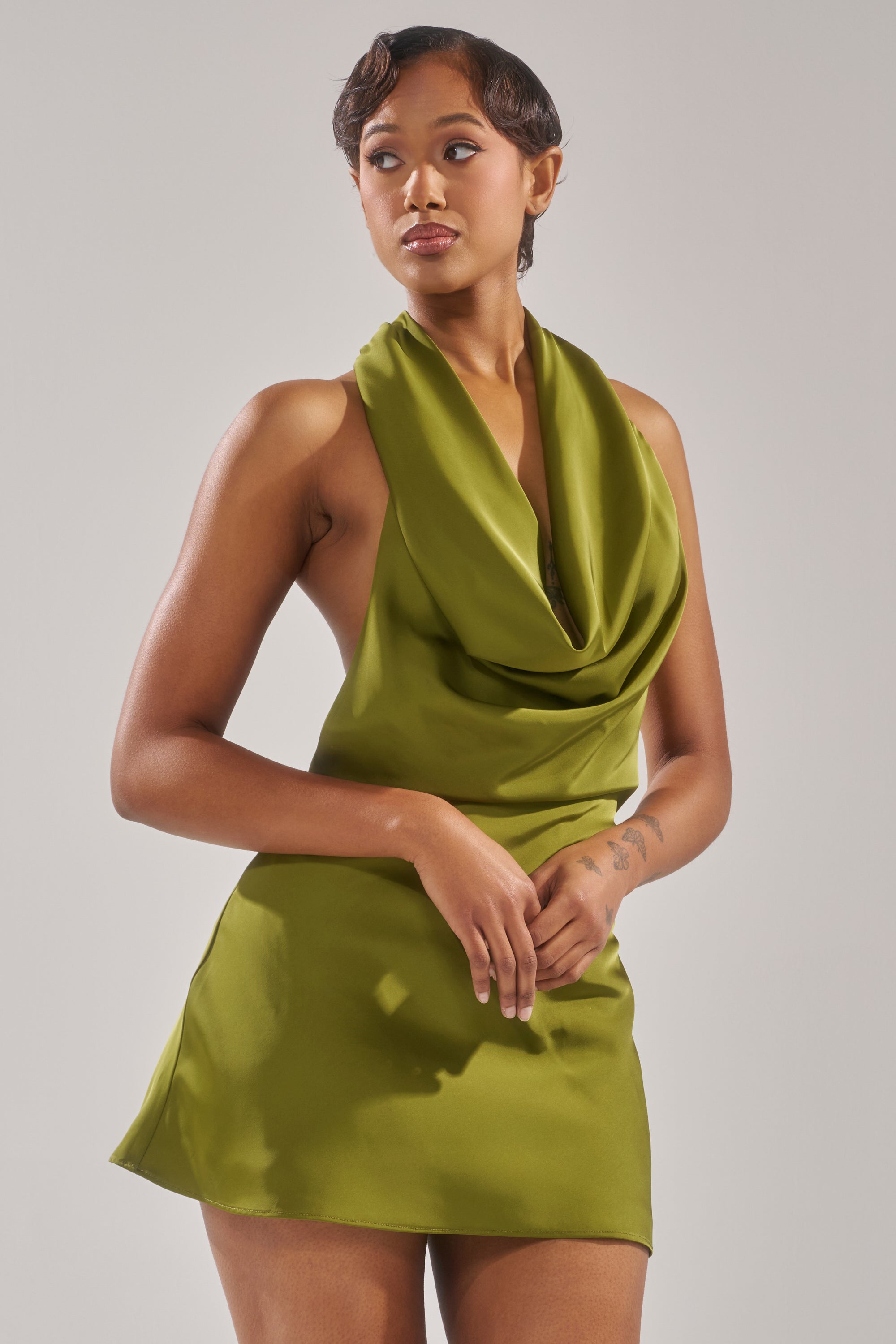 A person stands against a plain background wearing the GIGI MINI DRESS IN GREEN. Their hair is short and styled, expression neutral, with hands resting in front of their body.
