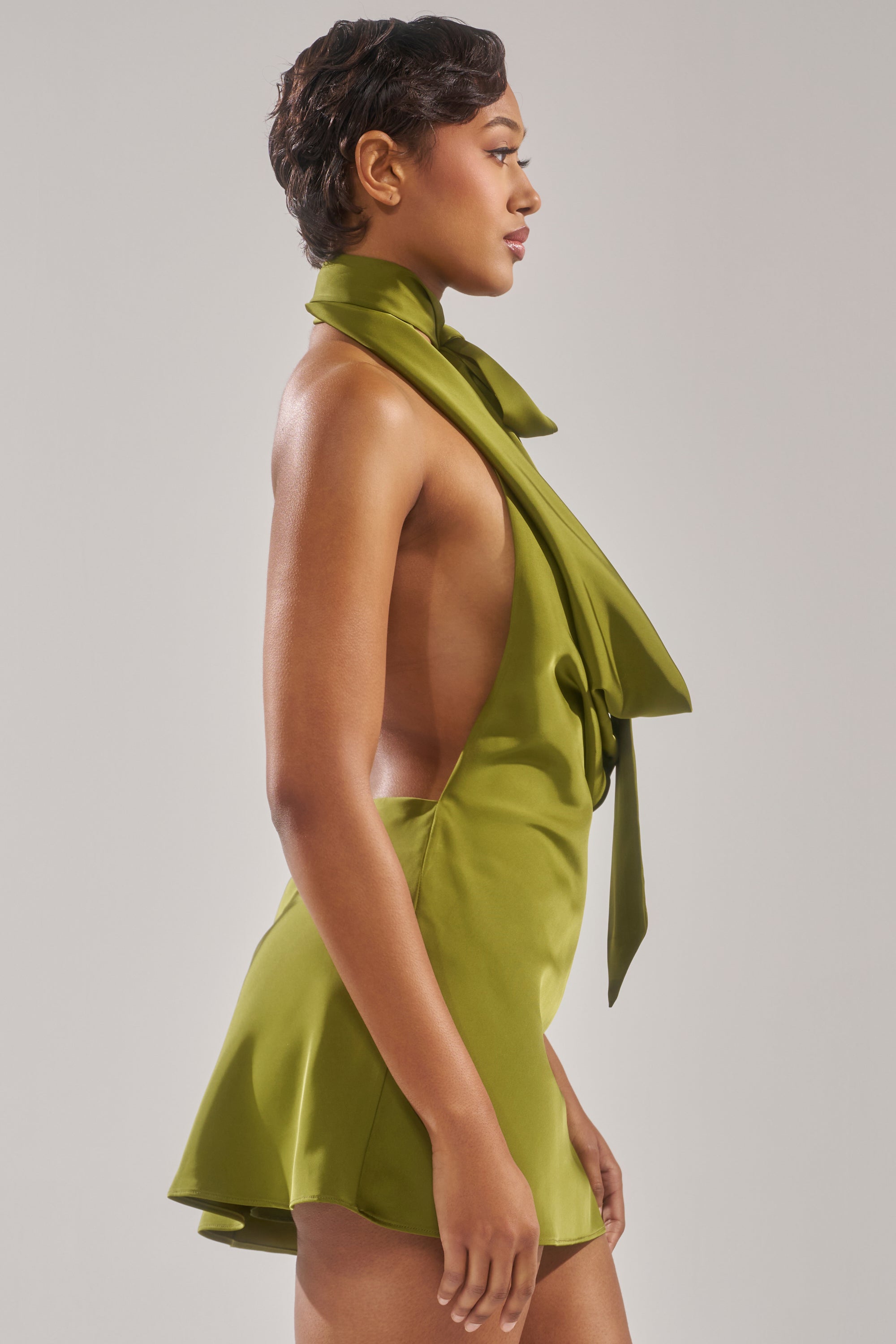 A woman in profile wears the GIGI MINI DRESS IN GREEN, a sleeveless, open-sided dress with a scarf-like neck detail, against a plain light gray background.